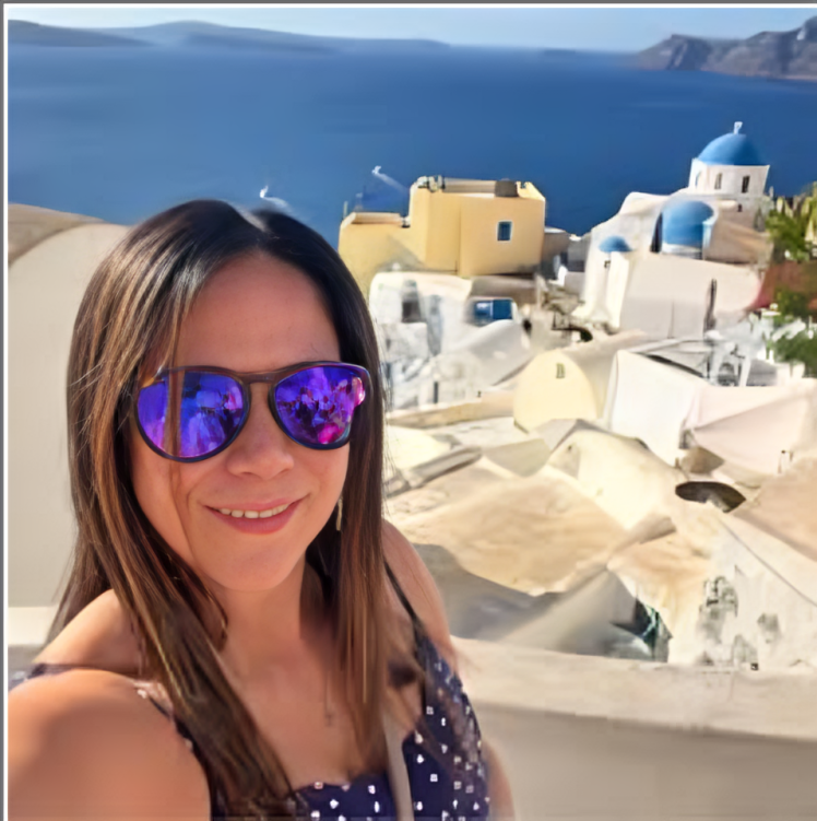 Woman wearing sunglasses smiles in front of a whitewashed village in Santorini, Greece. The blue Aegean Sea is in the background.