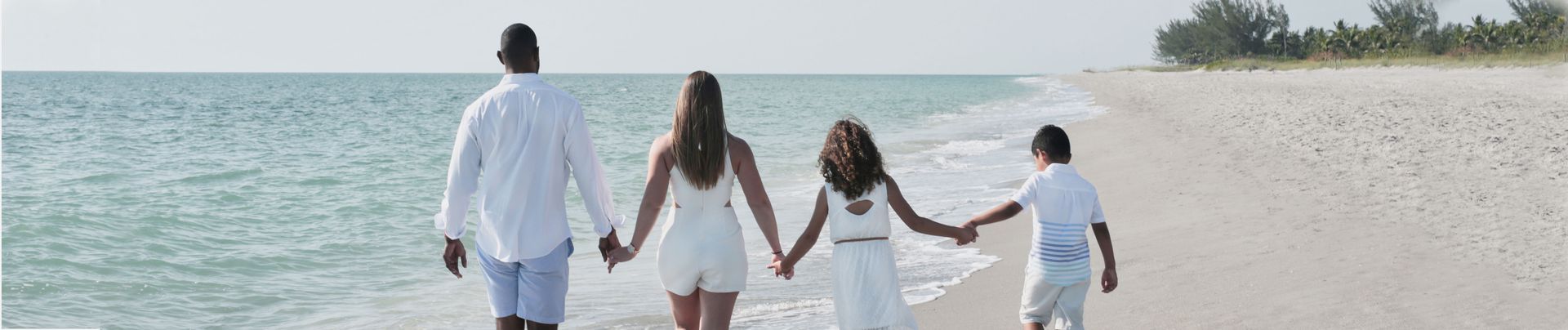 A family of four walks hand-in-hand along a sandy beach. They are dressed in white and facing away from the viewer, towards the ocean.