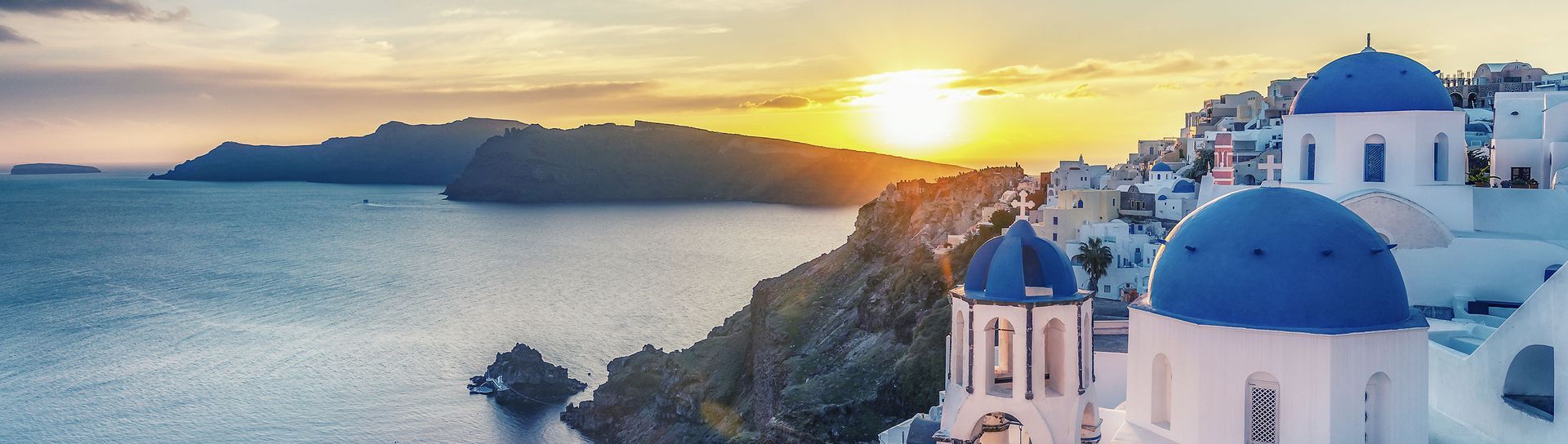 Sunset over the Aegean Sea, Santorini, Greece; white buildings with blue domes, coastline, and the sun setting on the horizon.