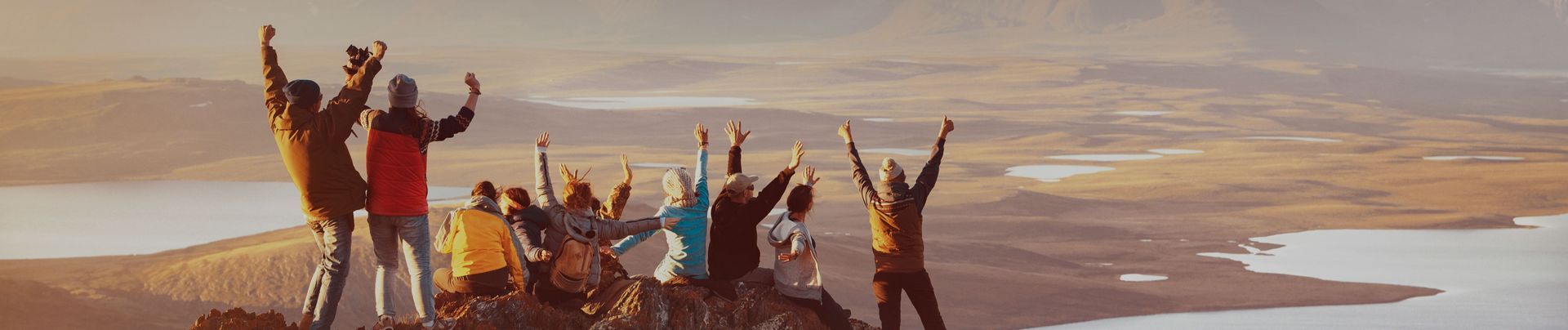 A group of people celebrating on a mountain peak, arms raised. The background features a vast landscape and a body of water.