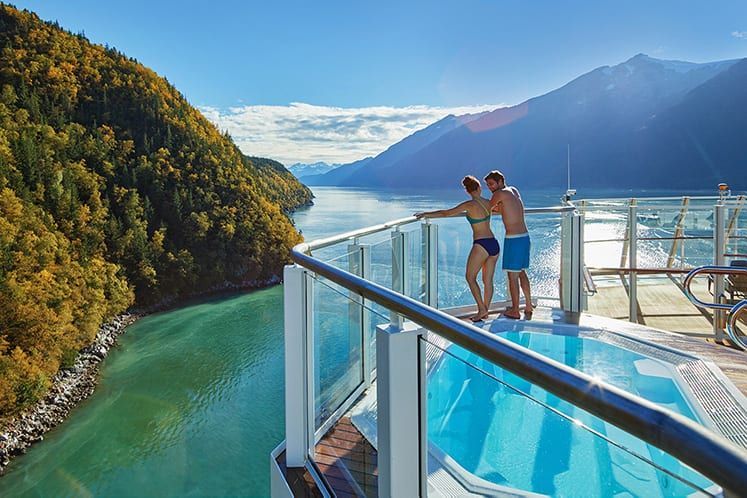A couple stands by a hot tub on a ship deck, overlooking a fjord with autumn trees and mountains.