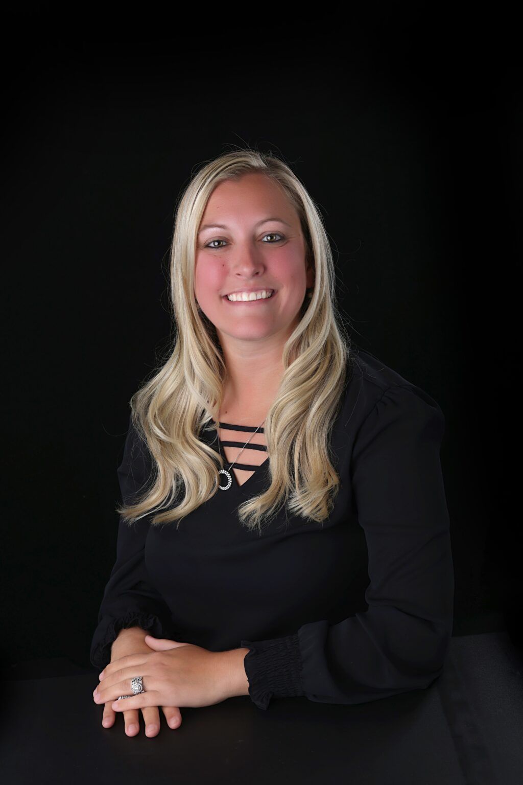 Woman with blonde hair smiles while looking at the camera. She wears a black top and sits against a black background.