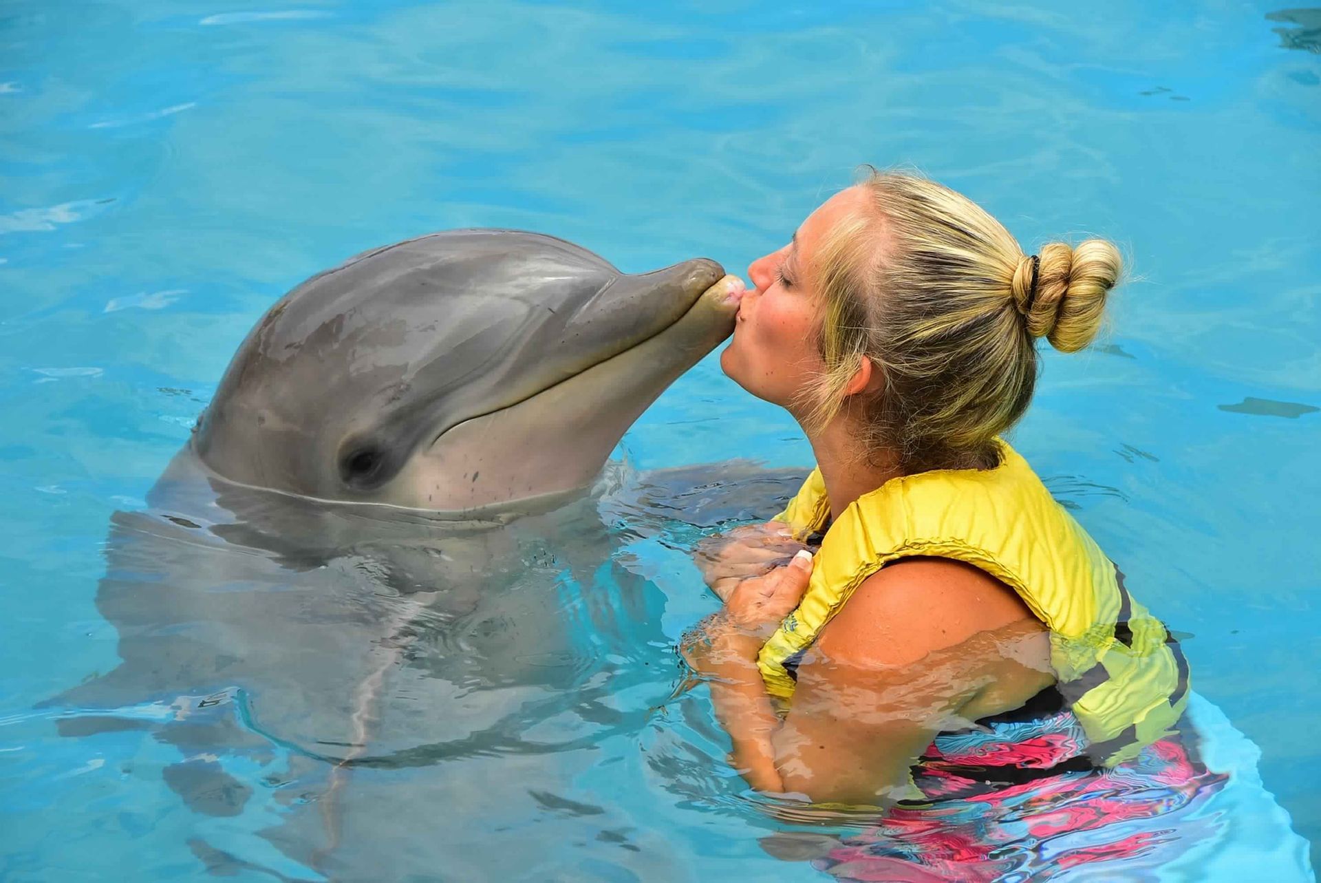 Woman in a life vest kissing a dolphin in a clear blue pool. The dolphin is gray and facing her.