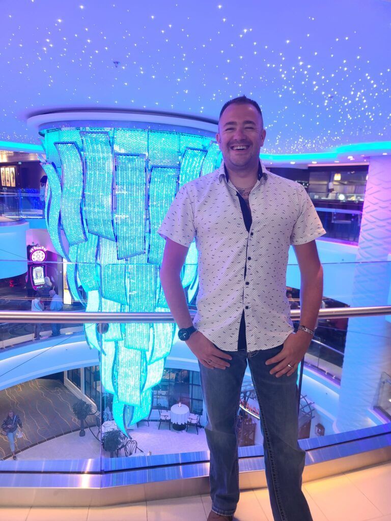 A smiling man poses on a balcony with a large, blue chandelier in the background. He's wearing a white shirt, jeans, and a watch.