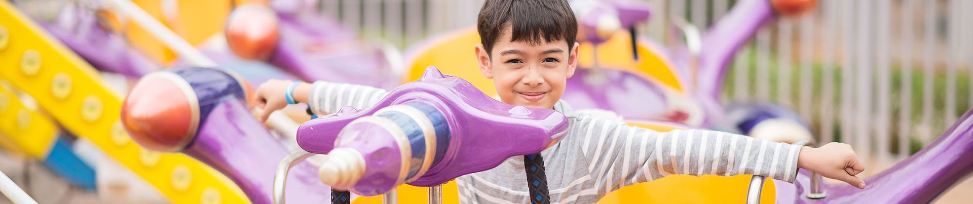 A young boy smiles, arms outstretched while riding an amusement park ride shaped like purple airplanes.