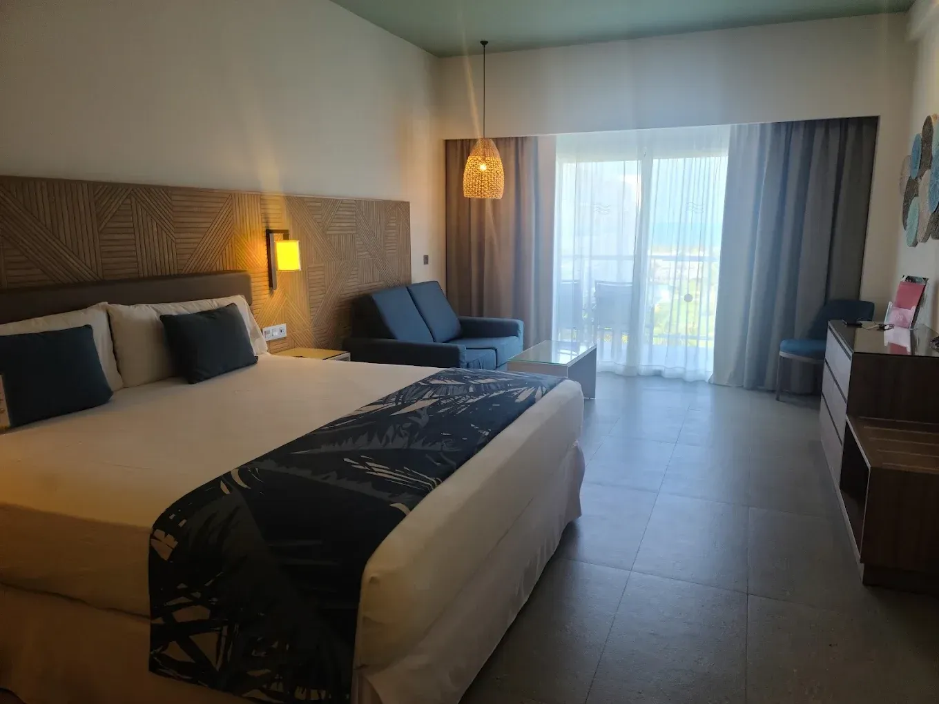 Hotel room with a king-sized bed, couch, and balcony access. The decor is neutral with a tropical vibe.