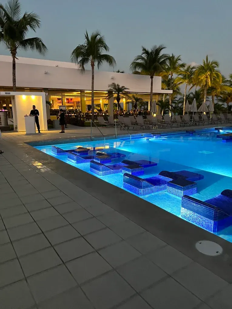 Pool with blue illuminated in-water seating at dusk, palm trees, and a building with dining area. A person stands at a bar.