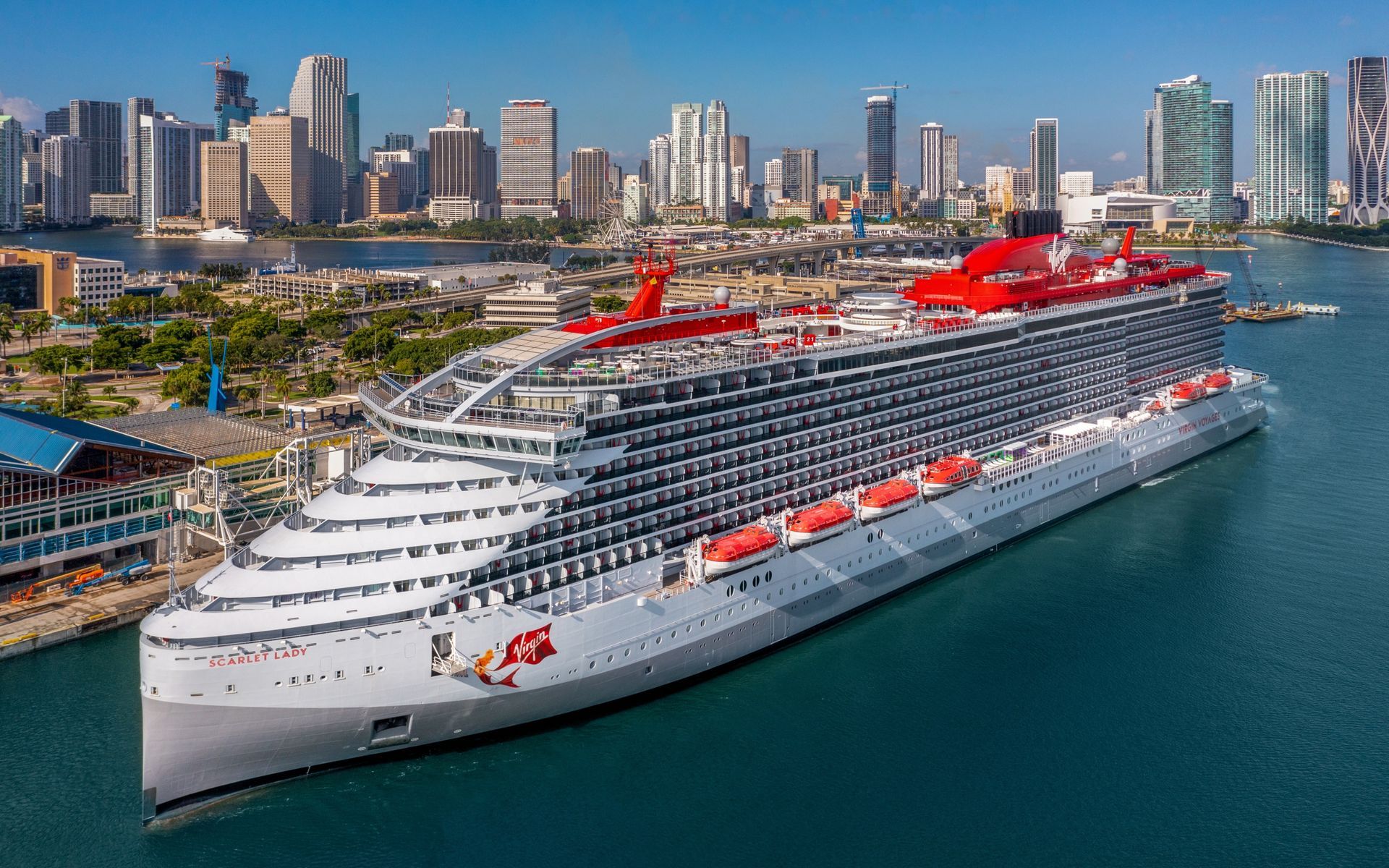 A large, gray and red cruise ship docked in a harbor, with a city skyline in the background.