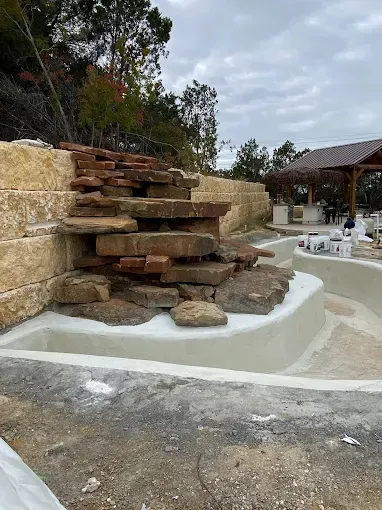 A tiered stone waterfall feature under construction next to a pool basin and a wooden pavilion under a cloudy sky.