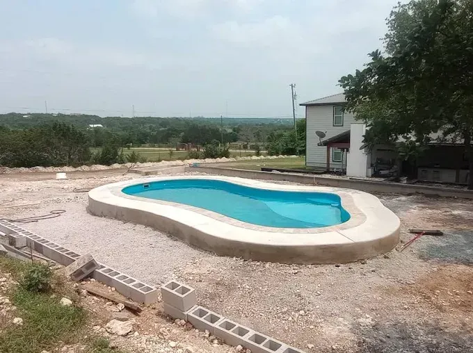 A freeform turquoise swimming pool with a concrete edge sits in a yard under construction, with a house in the background.