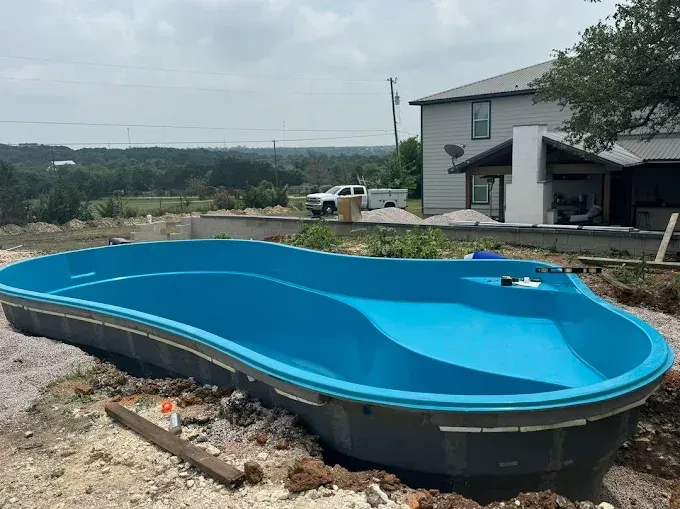 A new, bright blue fiberglass pool shell sits in a partially excavated backyard next to a house under construction.
