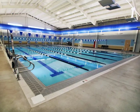 Indoor swimming pool with several lanes, blue lane markers, and a surrounding concrete deck under a high white ceiling.