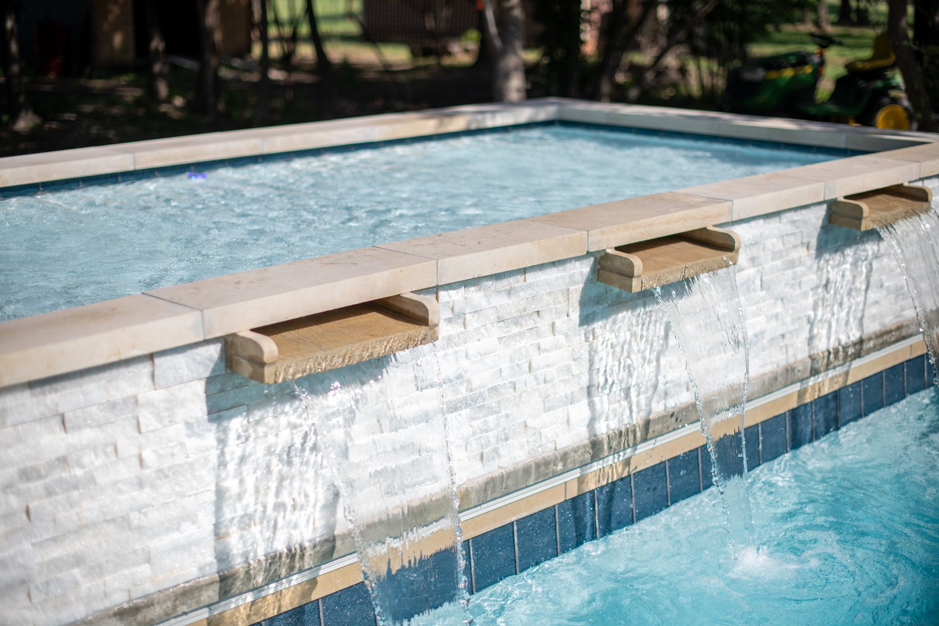 A white tiled raised spa wall with three stone scuppers pouring water into a swimming pool below.