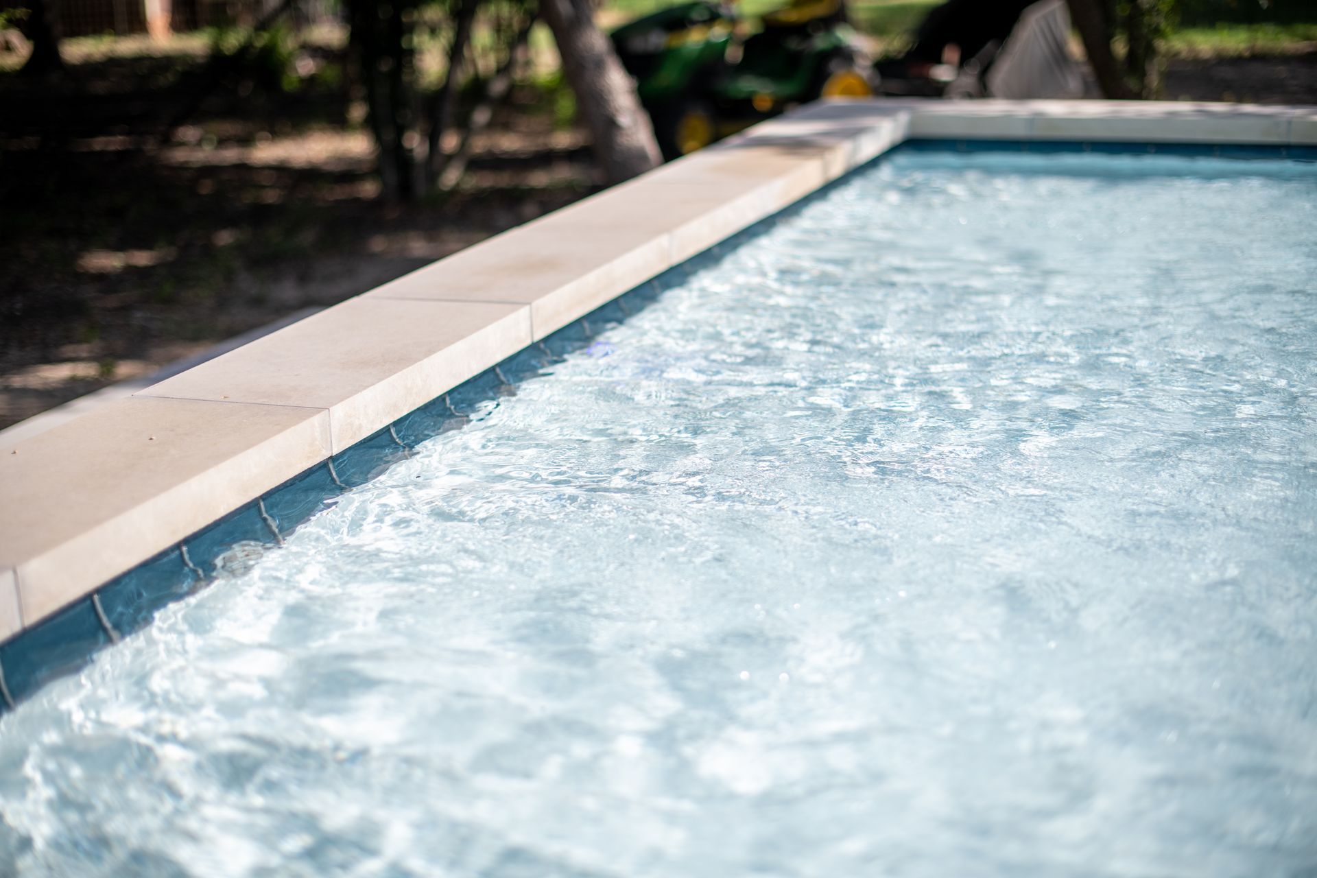 A close-up view of clear, rippling blue water in a swimming pool with light-colored stone coping along the edge.