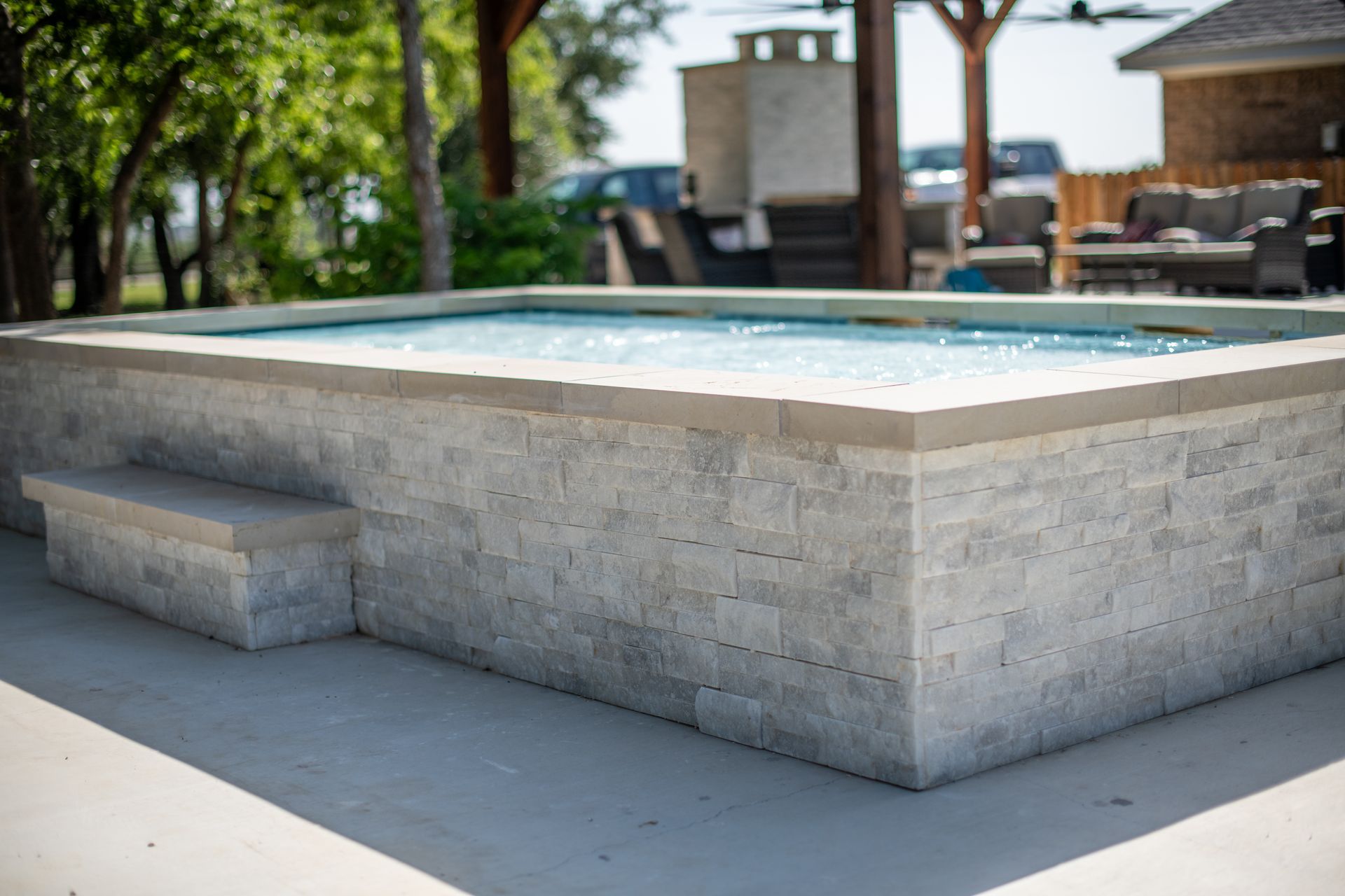 A raised, rectangular stone hot tub or plunge pool with a concrete step, situated on a patio near outdoor furniture.