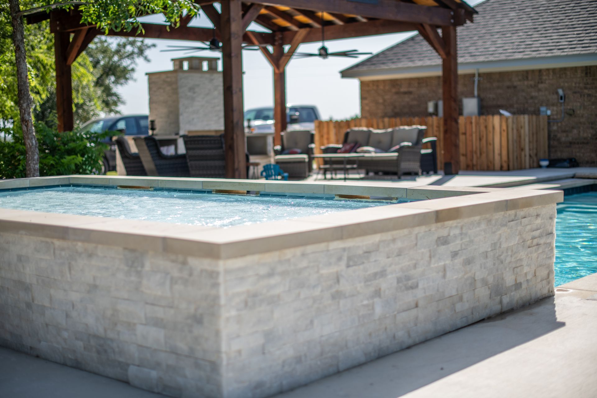A light-colored stone-faced raised hot tub adjacent to a swimming pool under a wooden pergola with patio seating.