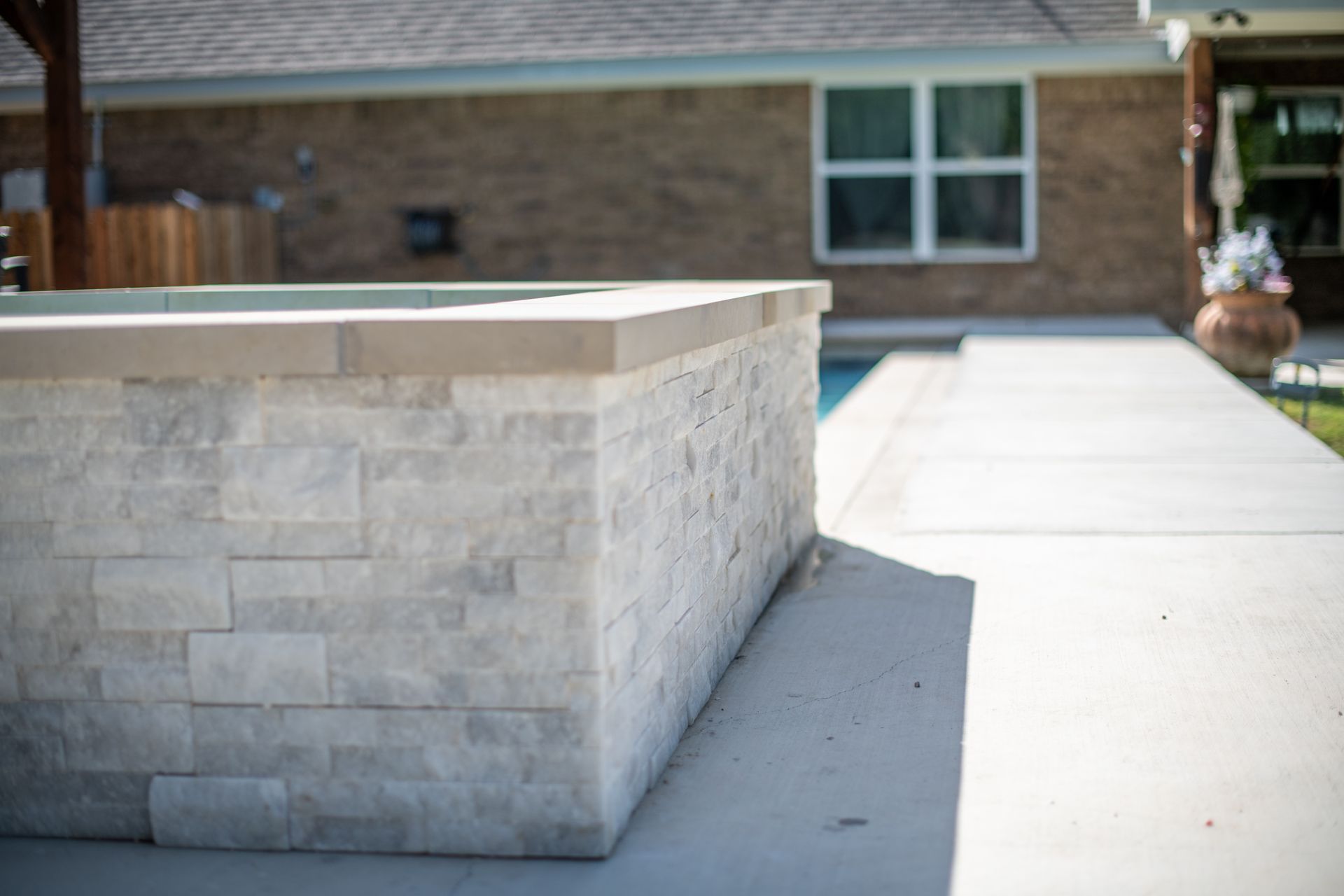 A close-up of a white stacked-stone outdoor fire pit beside a swimming pool, with a brick house in the background.