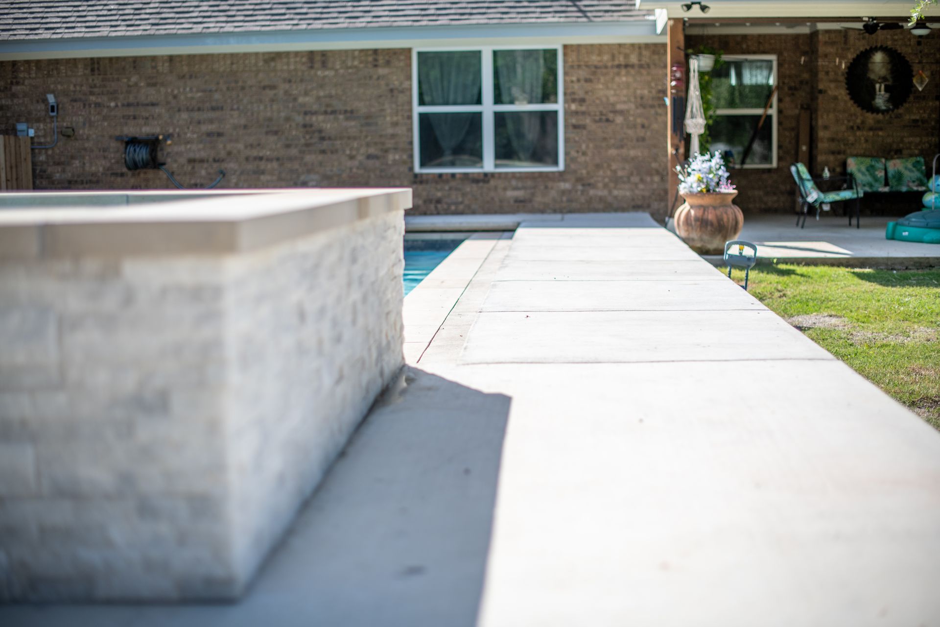 A raised stone pool feature with a smooth concrete deck in a residential backyard with a brick house.