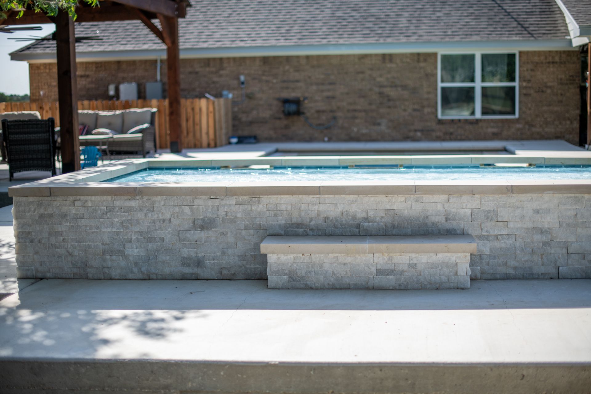 A raised pool with light gray stone siding and a stone bench, situated on a patio next to a house.