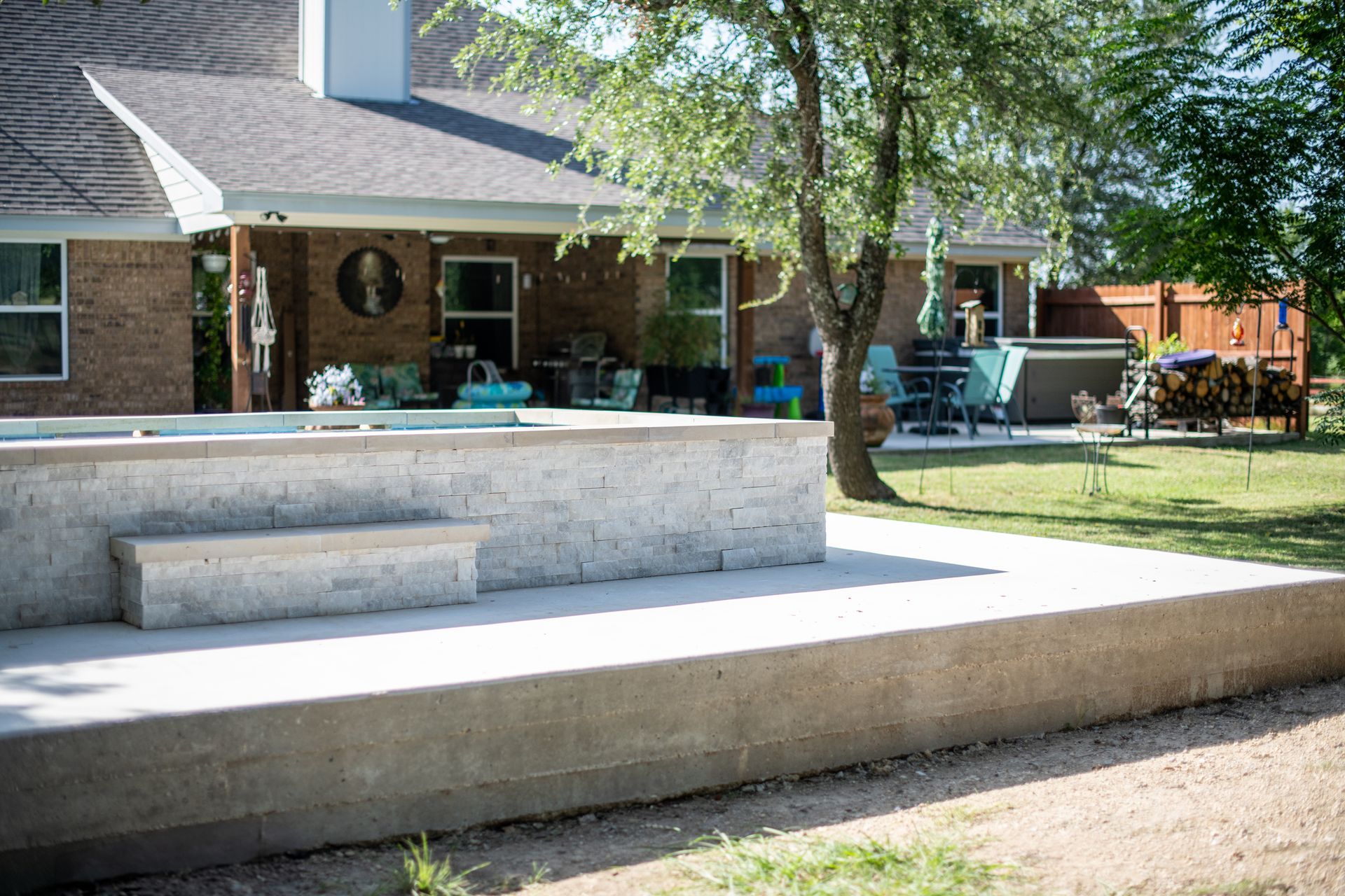 A raised stone patio and bench in a backyard, featuring a patio cover, outdoor furniture, and a tree on a sunny day.