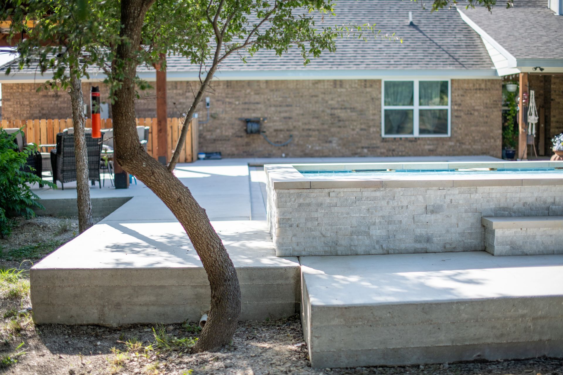 A raised, light-stone swimming pool and concrete patio situated in a backyard beside a brick house and a mature tree.