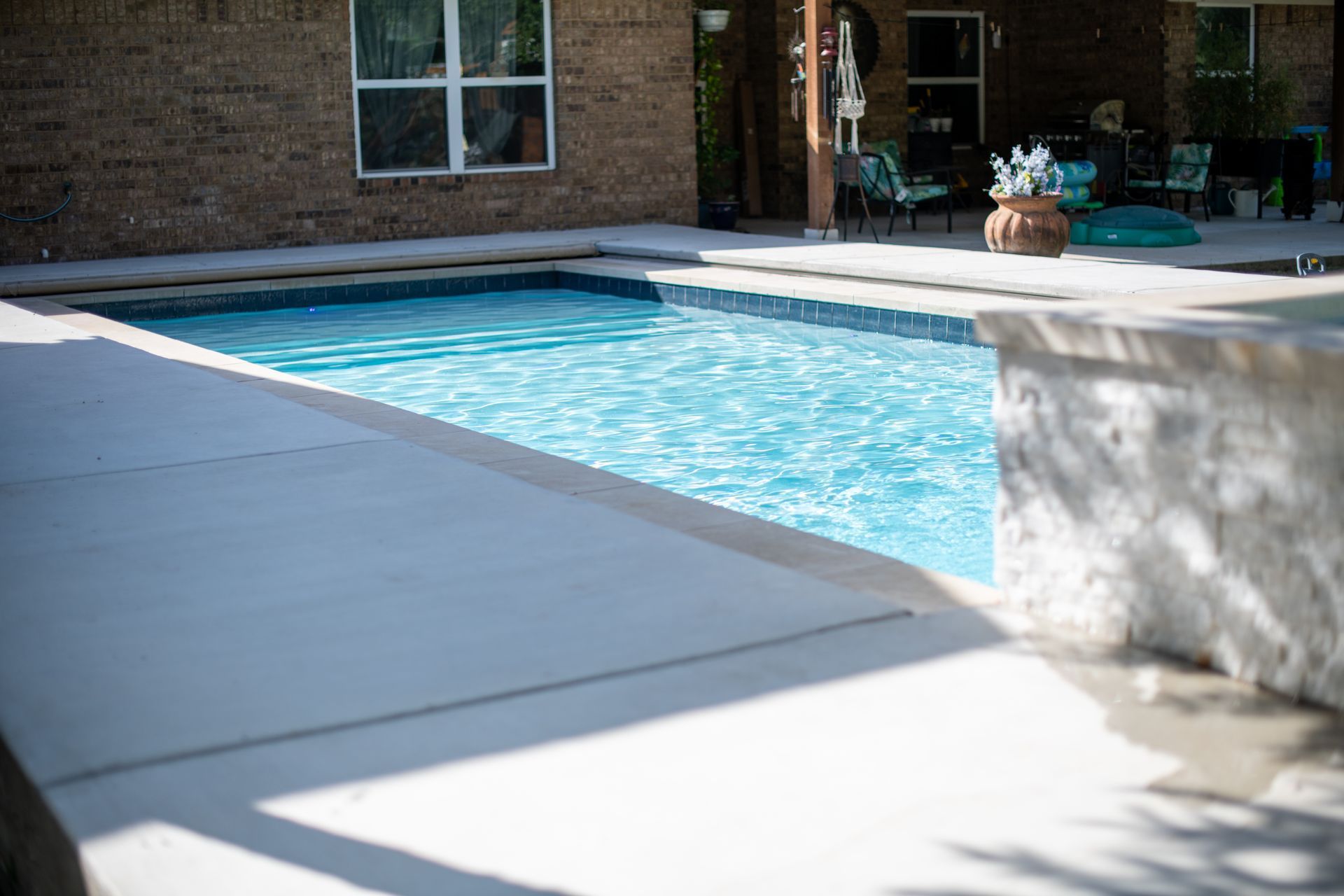 A clear, rectangular swimming pool surrounded by concrete decking and a stone wall, viewed from a patio in a backyard.