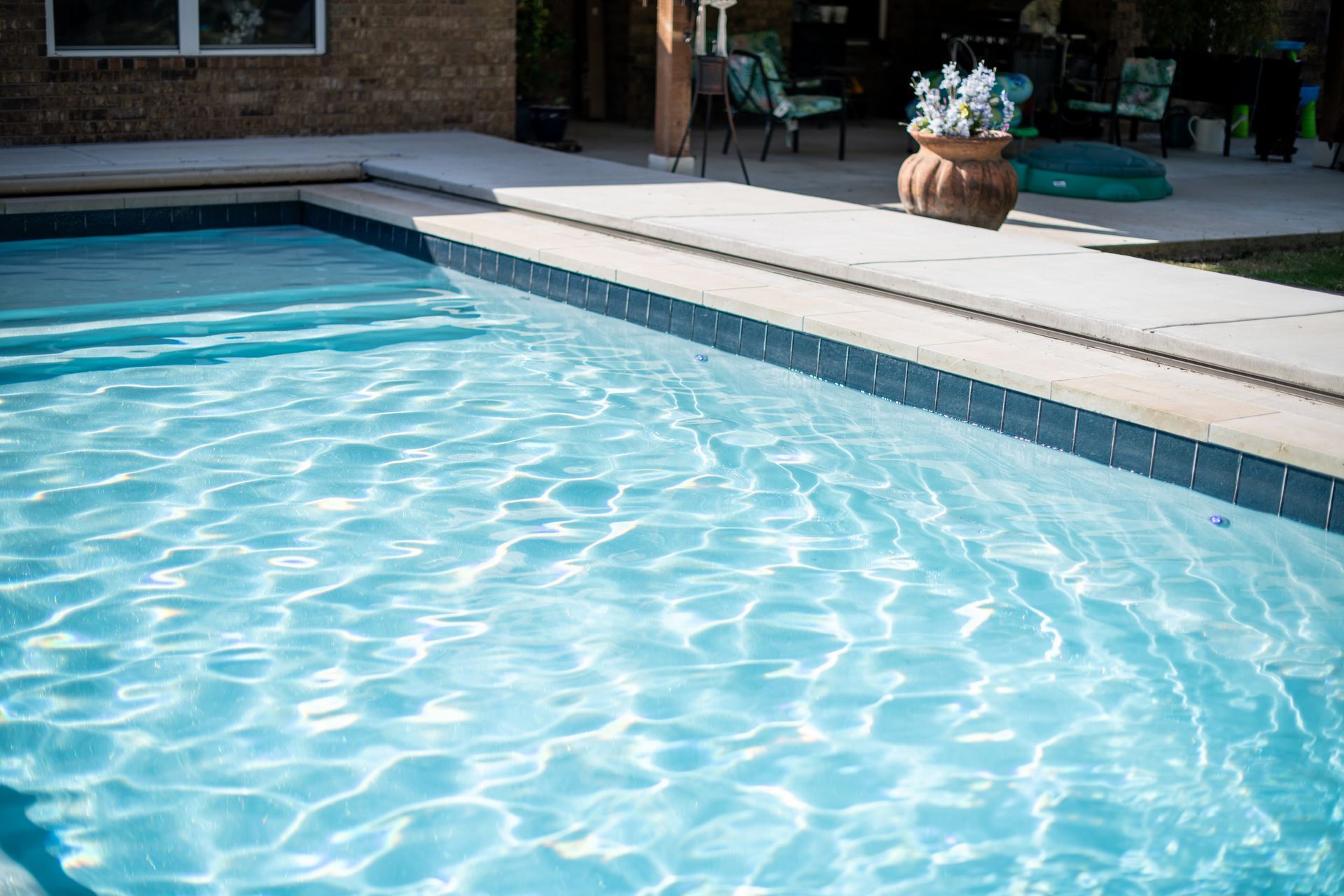 A backyard swimming pool with light blue rippling water, featuring stone coping and a paved patio area in the background.