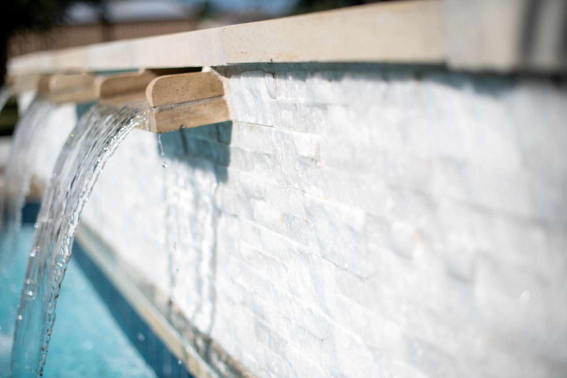 Water cascades from two small stone spouts into a turquoise swimming pool against a white, textured stone wall.