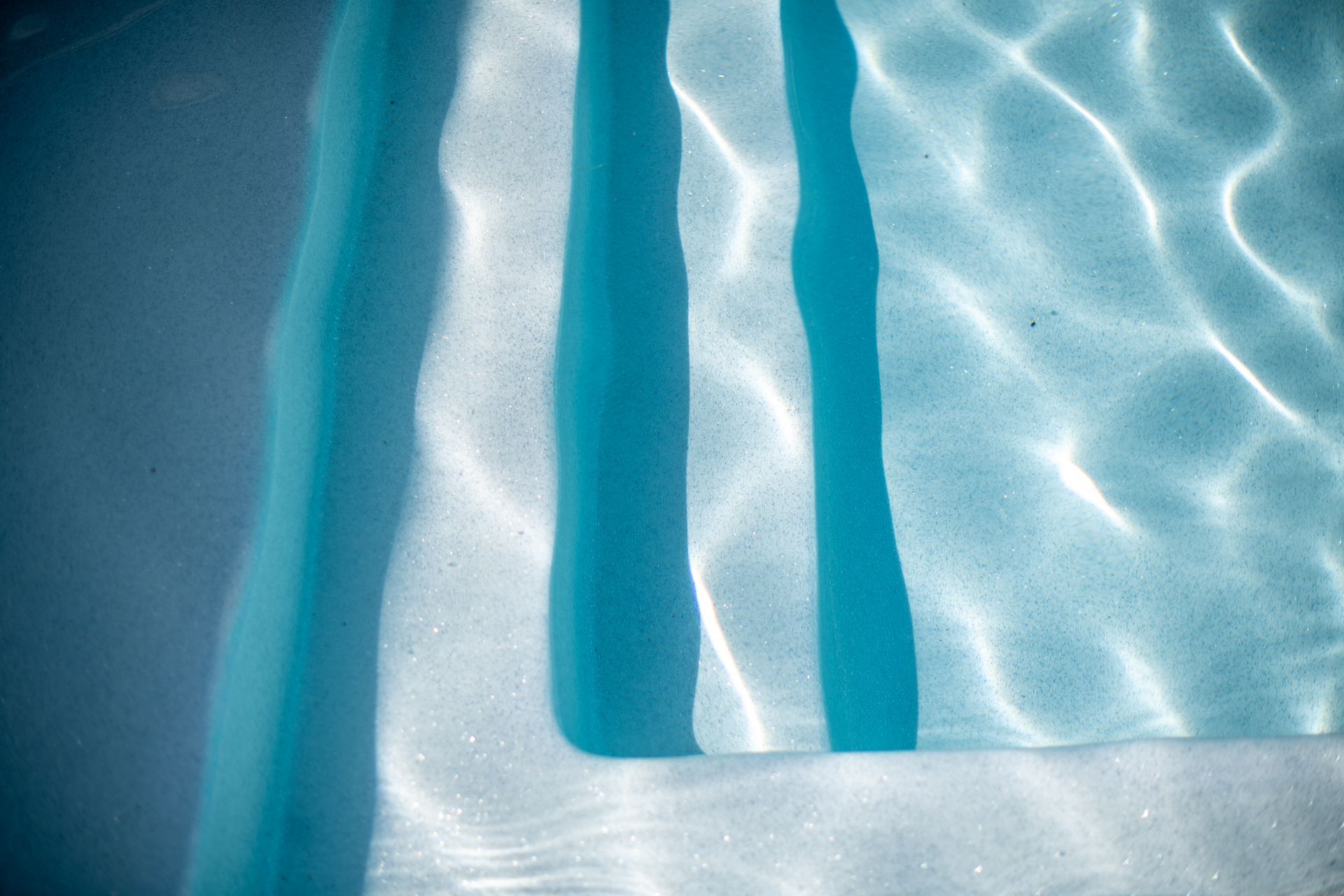 Light blue swimming pool stairs visible beneath clear, rippling water with sunlight reflections.