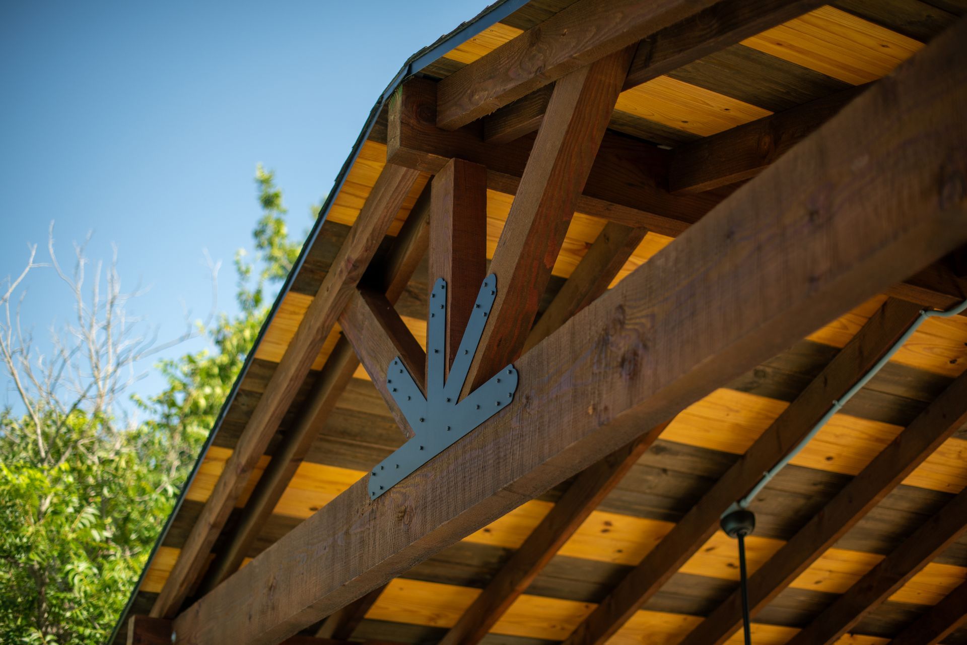 Close-up of a wooden timber roof truss with a decorative black metal plate, against a bright blue sky and green trees.