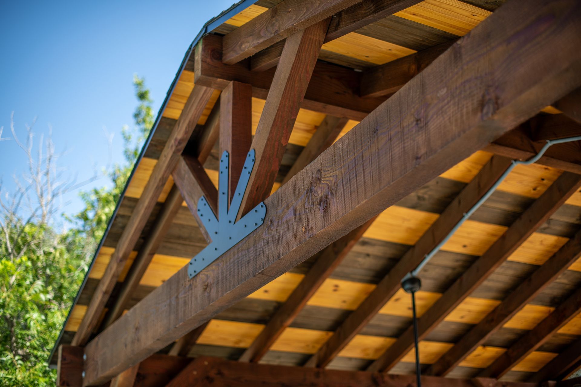 A wooden roof support structure features a decorative dark gray metal bracket connecting the beams, set against blue sky.