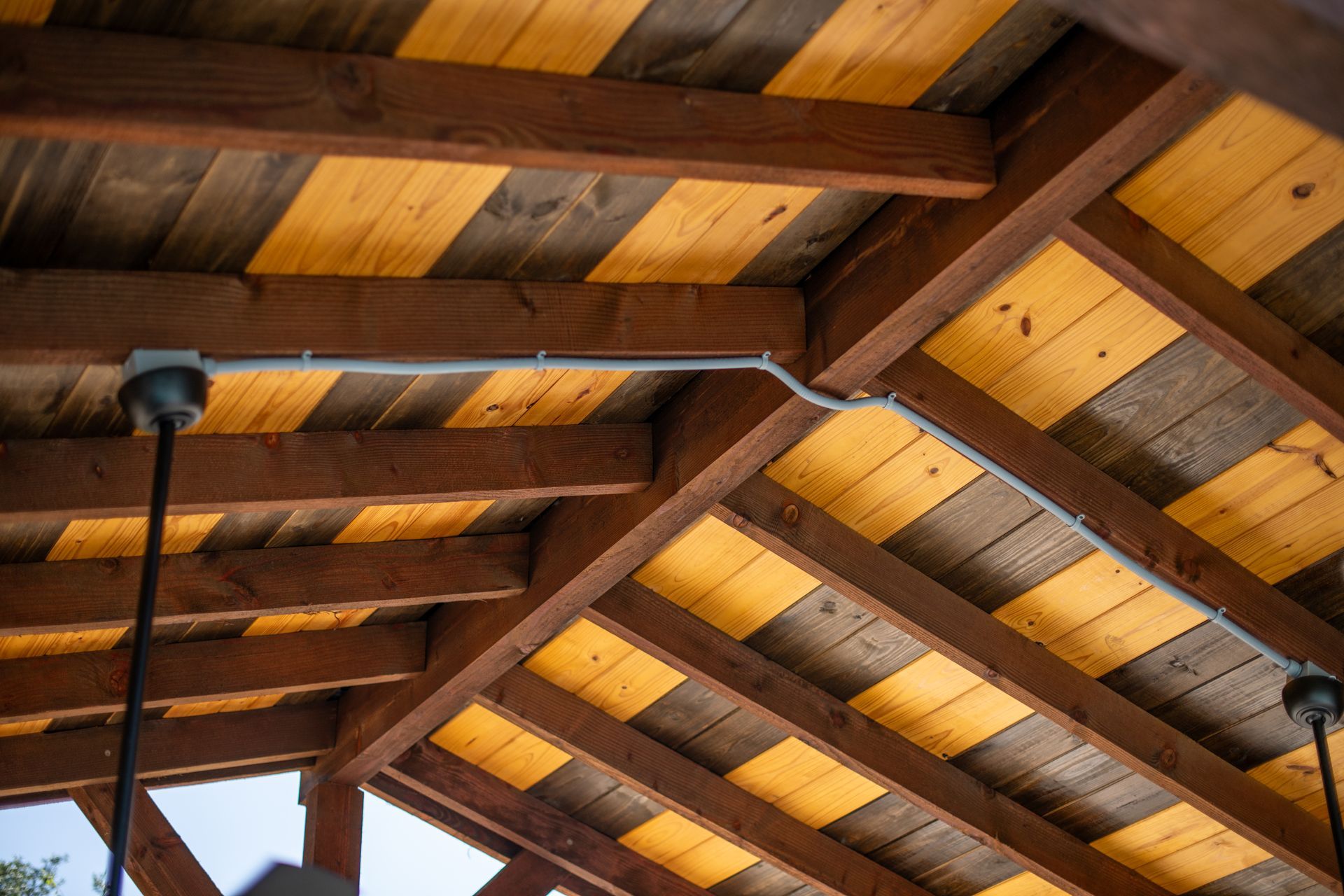 Wooden ceiling rafters with alternating light and dark planks, featuring exposed gray electrical conduit and light fixtures.