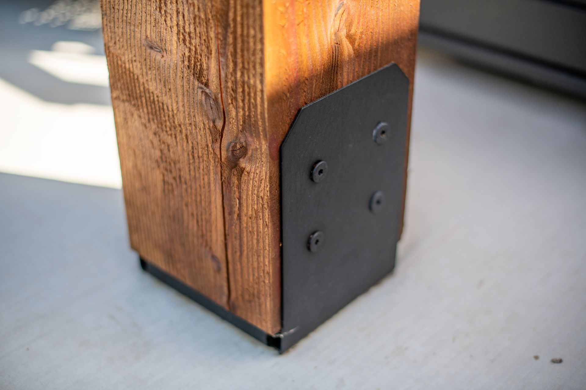 A stained wooden post secured to a concrete surface with a matte black metal base bracket and four visible screws.
