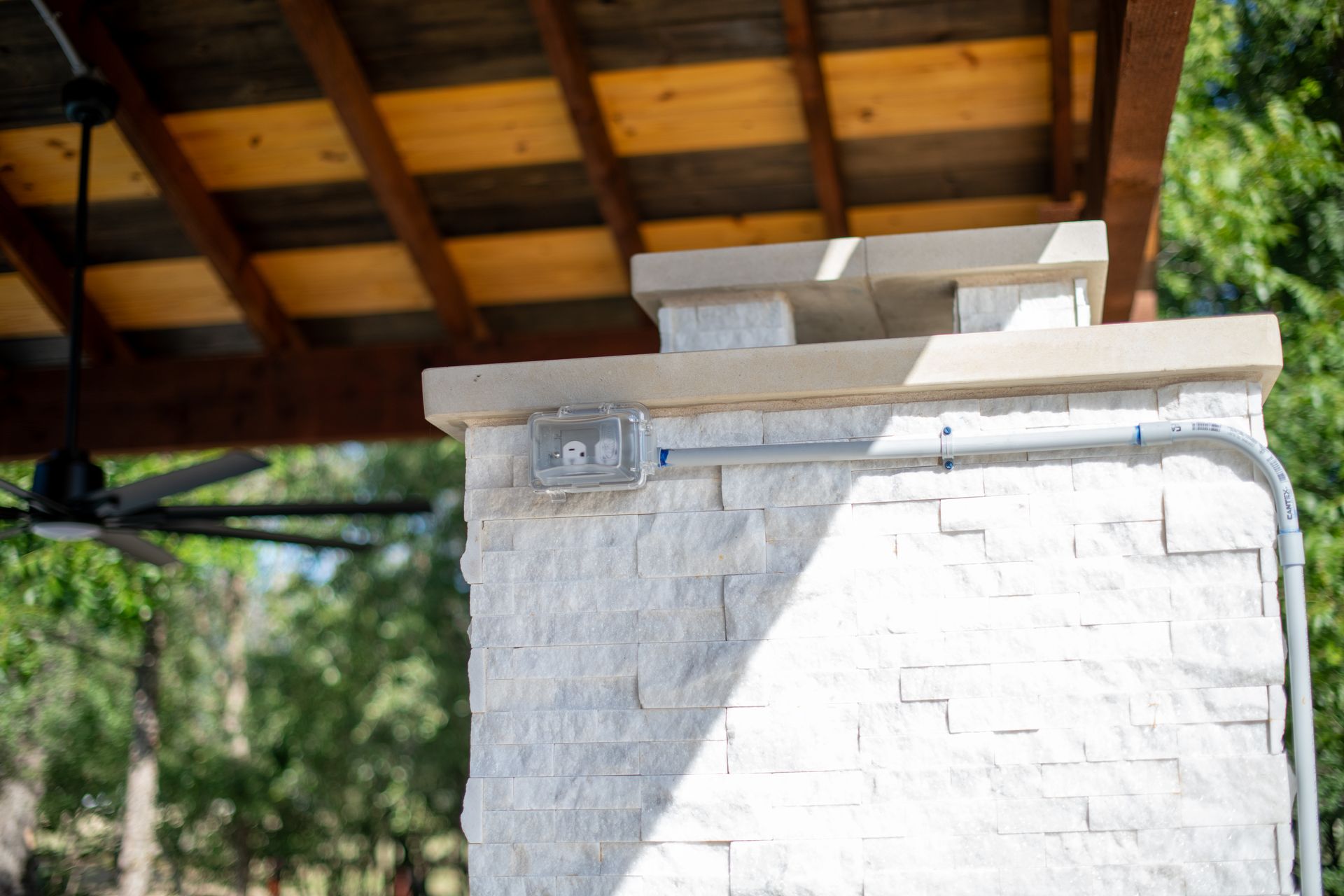 A stone pillar with an electrical outlet and conduit on an outdoor patio under a wooden pergola.