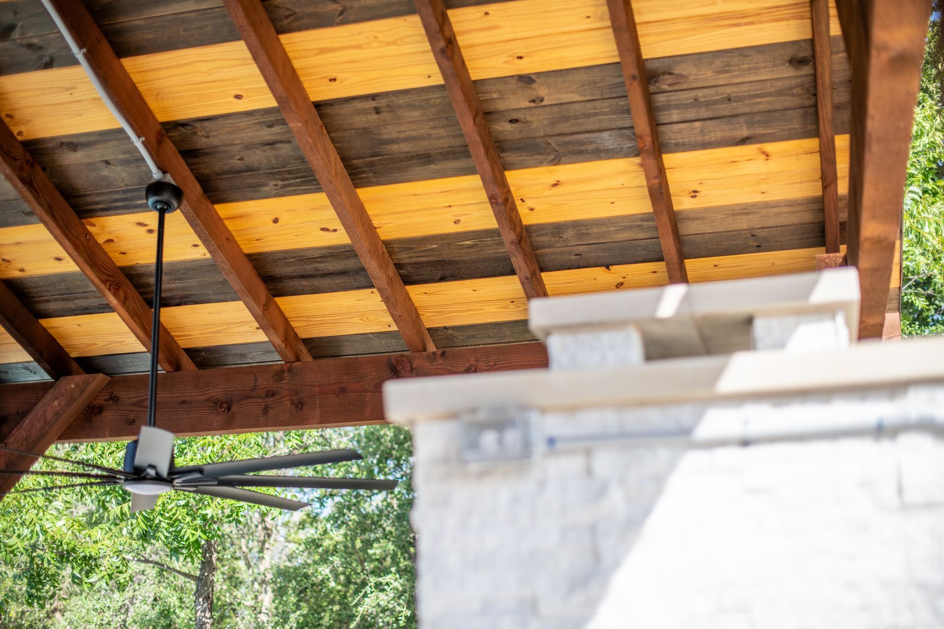 A low-angle view of a wooden pergola ceiling with alternating brown and yellow panels and a ceiling fan over a stone hearth.