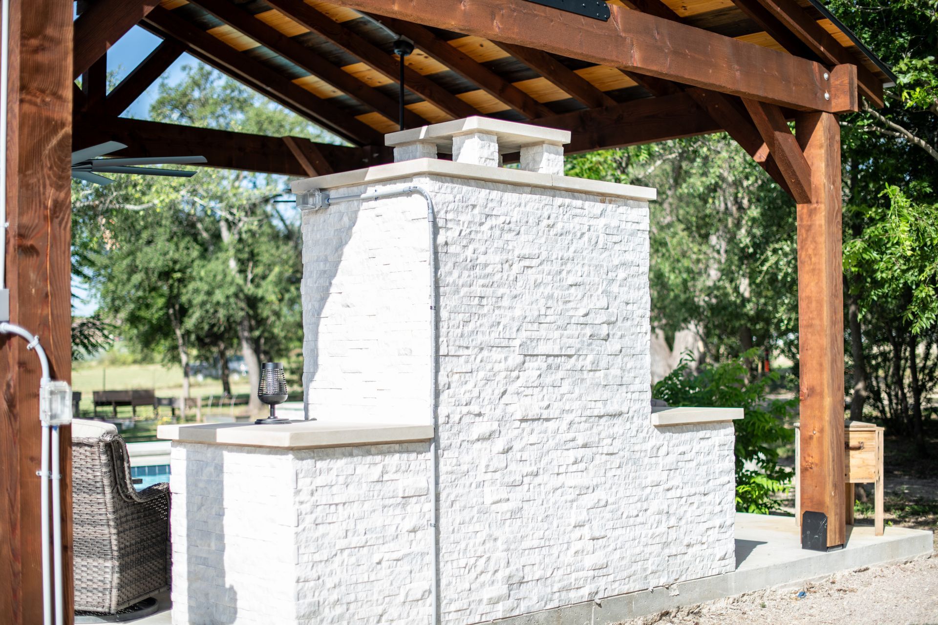 A white stone outdoor fireplace under a wooden patio cover in a backyard setting.