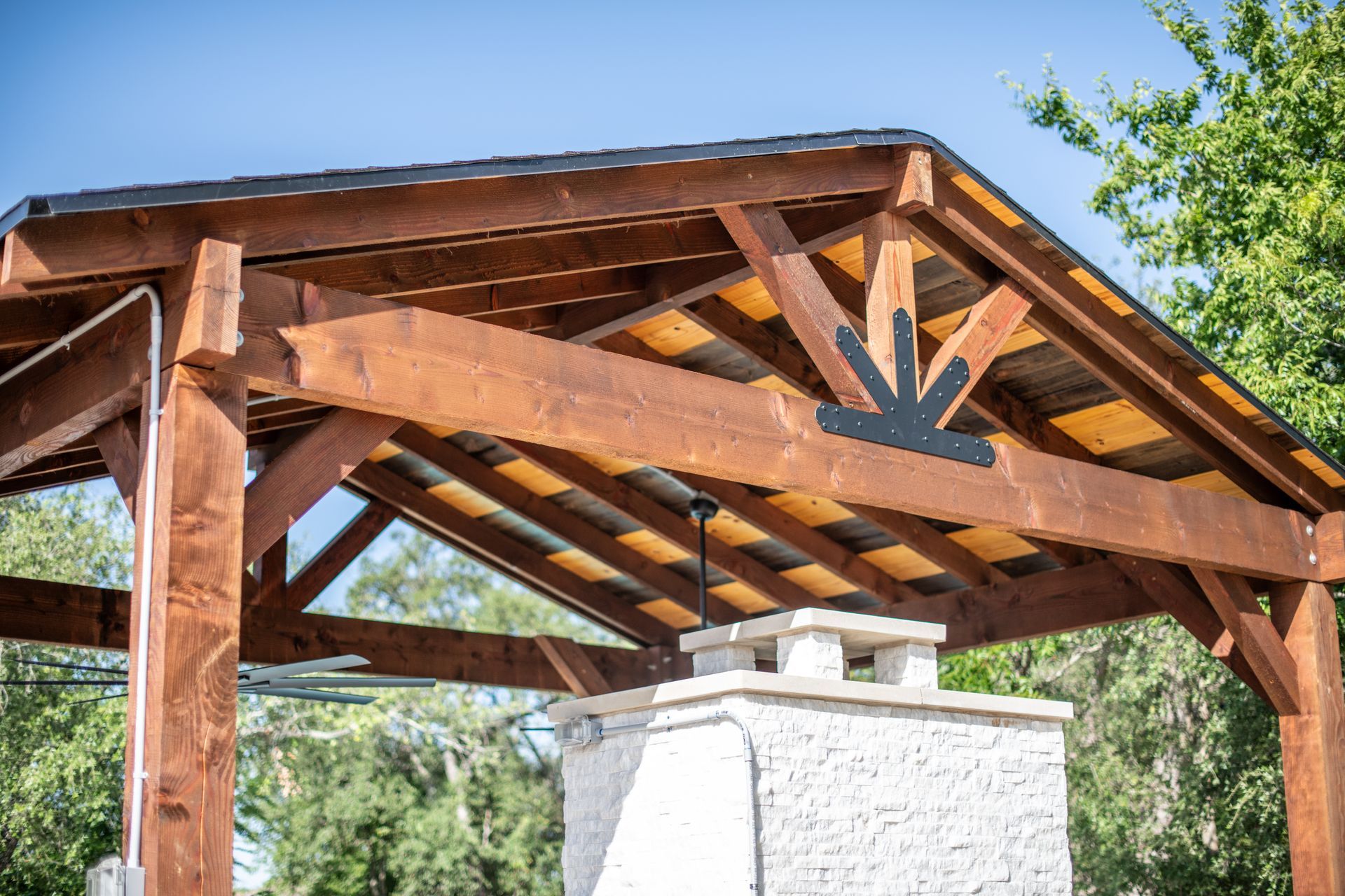 A wooden timber-framed patio cover with a black decorative bracket over a stone fireplace under a clear blue sky.