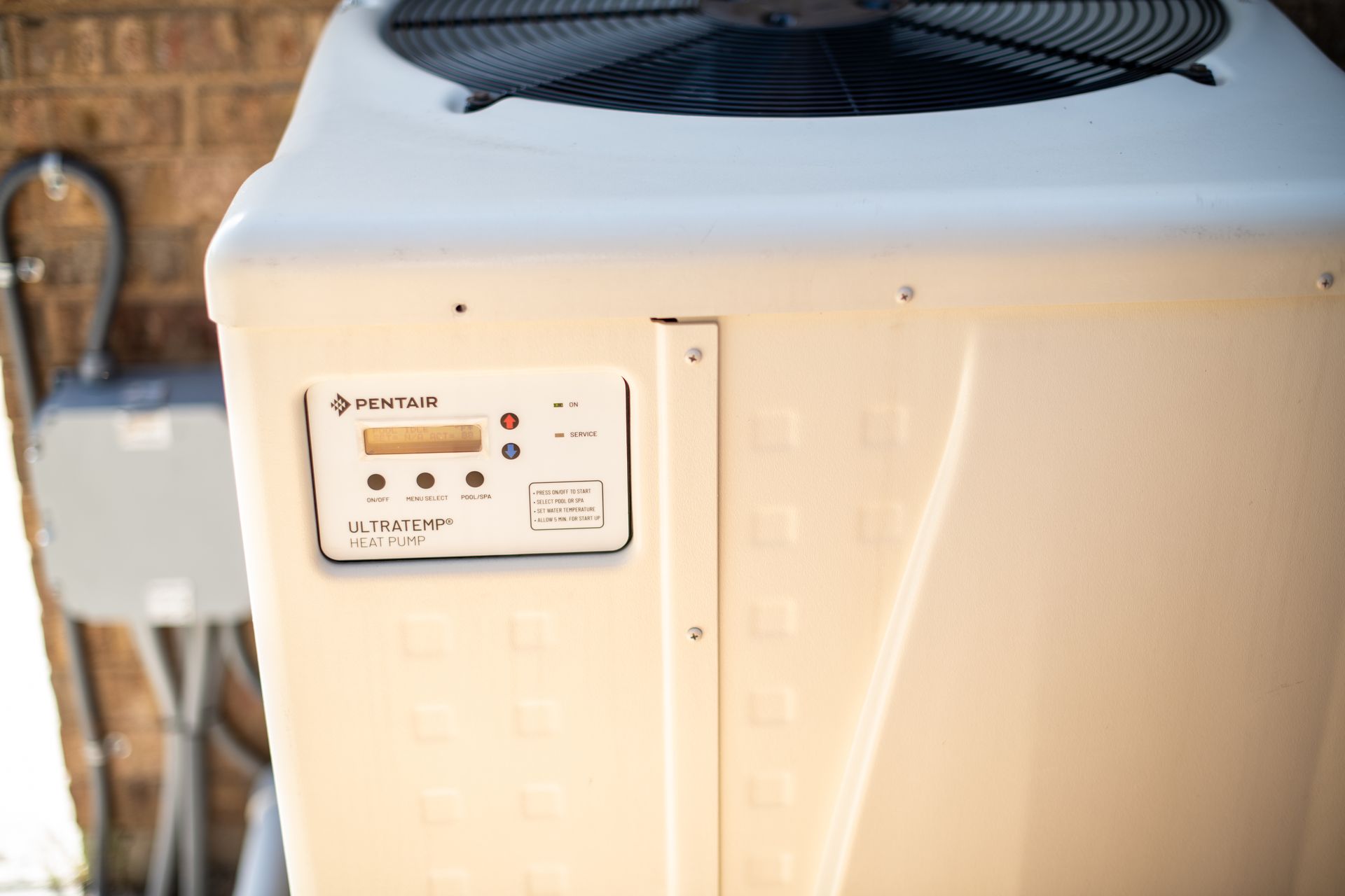 A white Pentair swimming pool heat pump with a digital control panel, situated outdoors next to a brick wall.