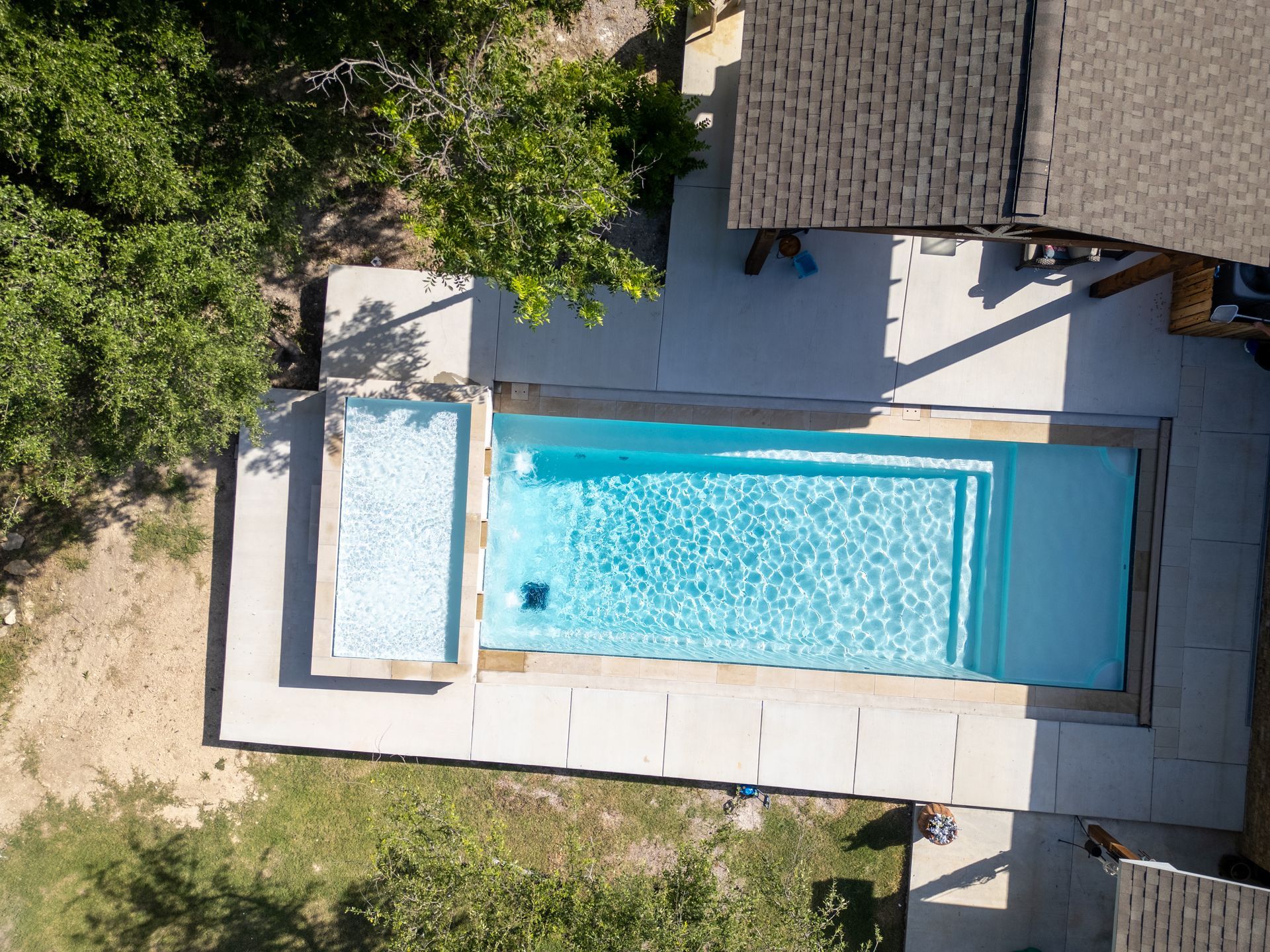 Aerial view of a rectangular blue swimming pool with an attached shallow spa area, surrounded by a light stone patio.