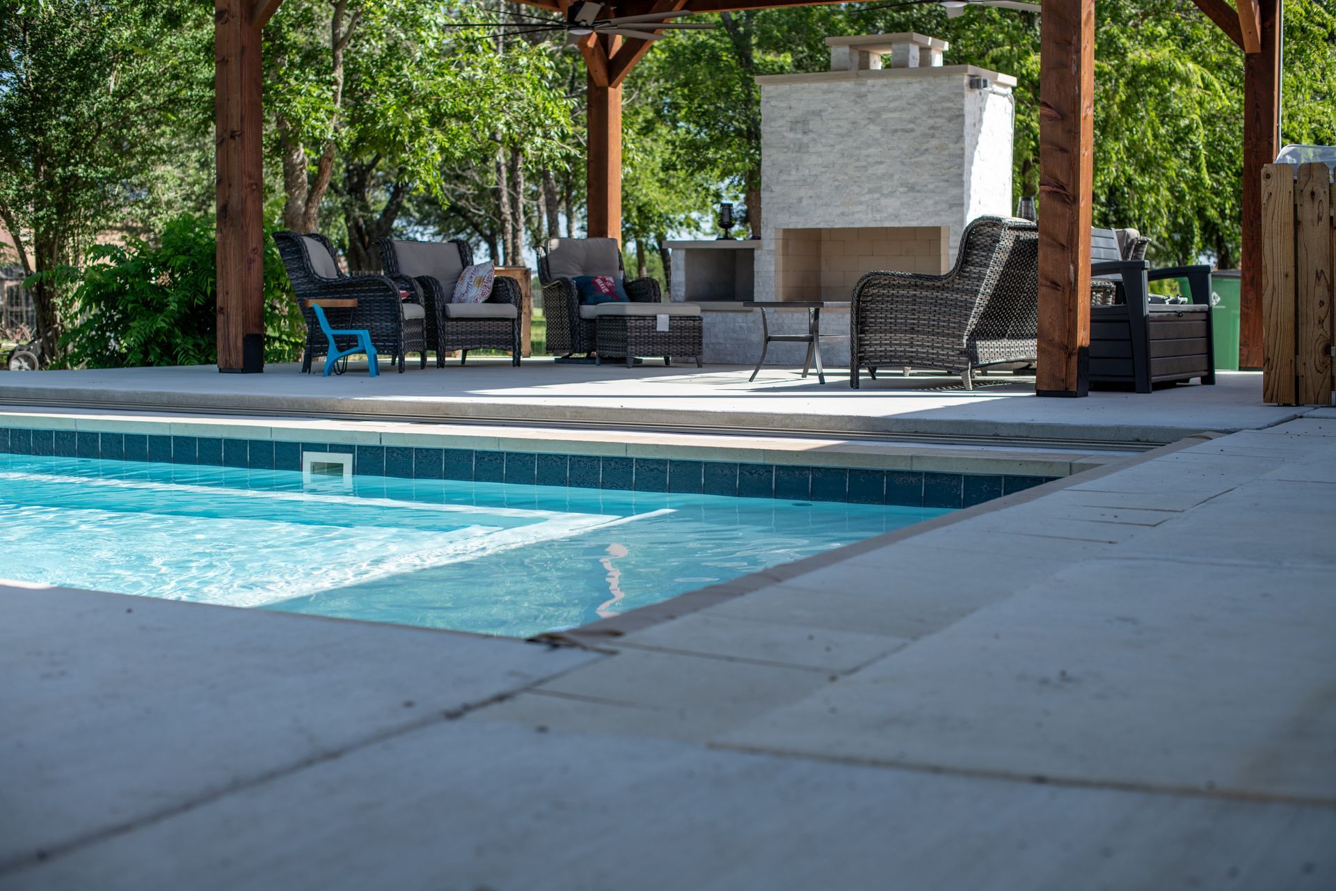 A swimming pool with blue water next to a covered outdoor patio area featuring wicker chairs and a stone fireplace.