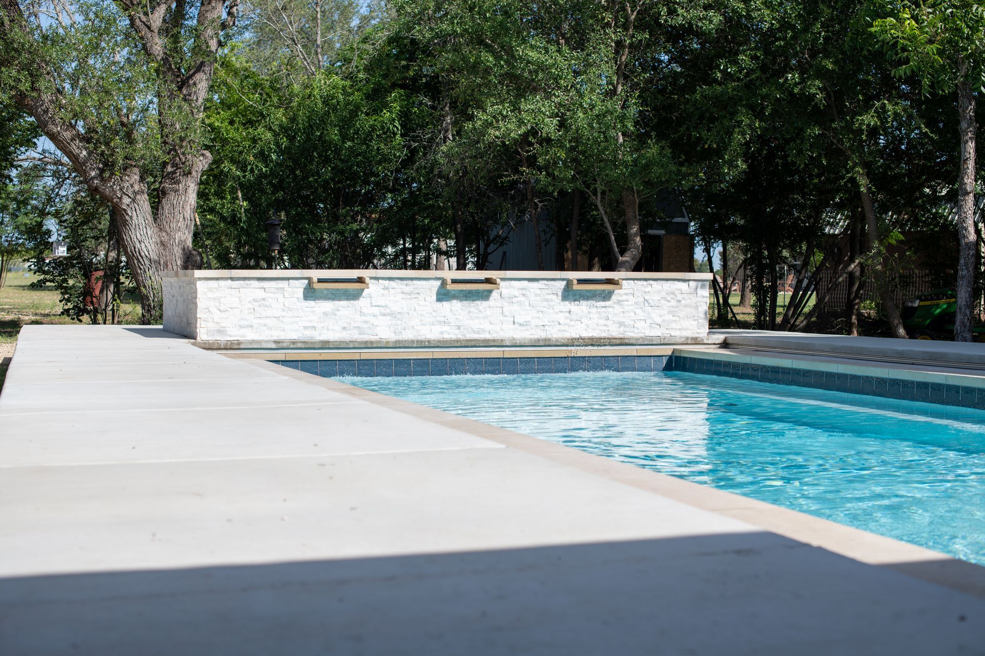 A bright blue backyard swimming pool with a white stone wall featuring water scuppers, set against a backdrop of trees.