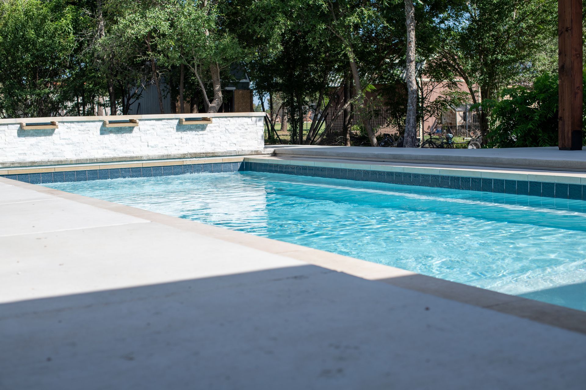 A rectangular swimming pool with blue water and a white stone wall with three water fountains in a sunny backyard setting.
