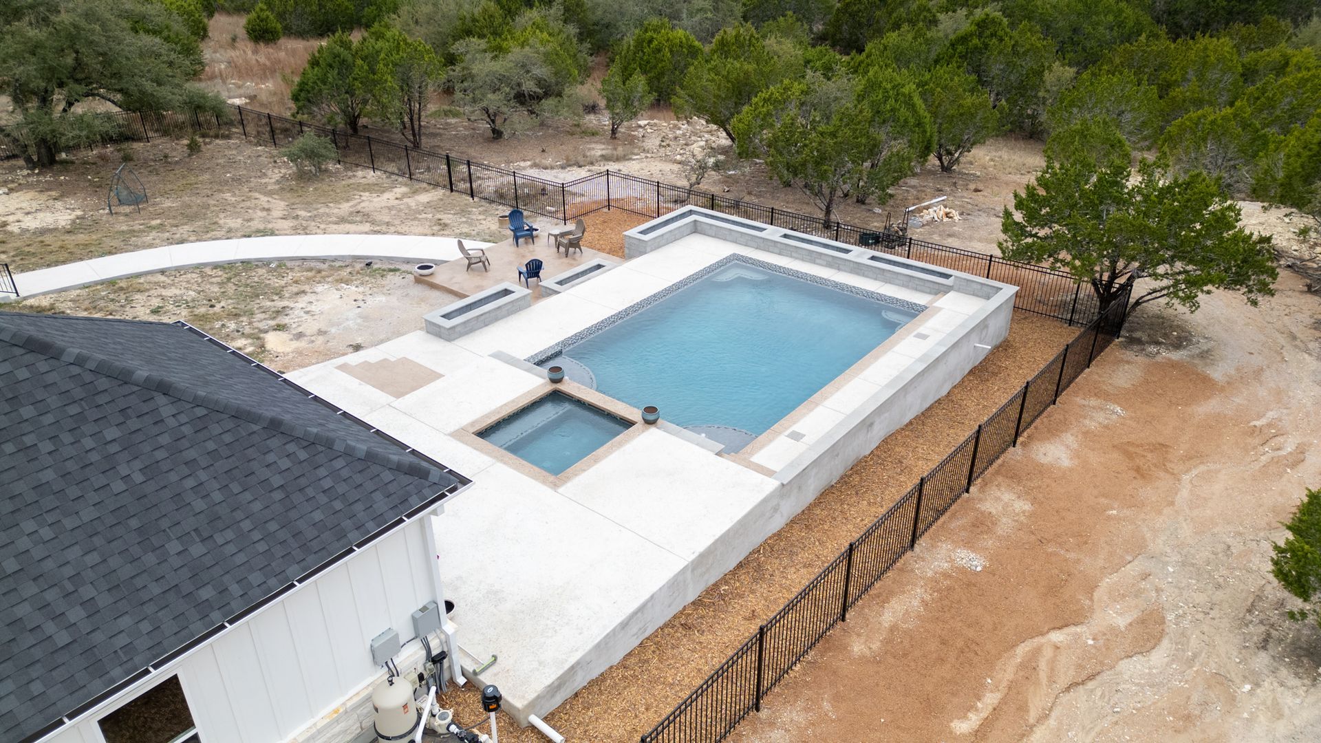An aerial view of a rectangular backyard pool with an integrated spa, set on a concrete patio next to a white house.