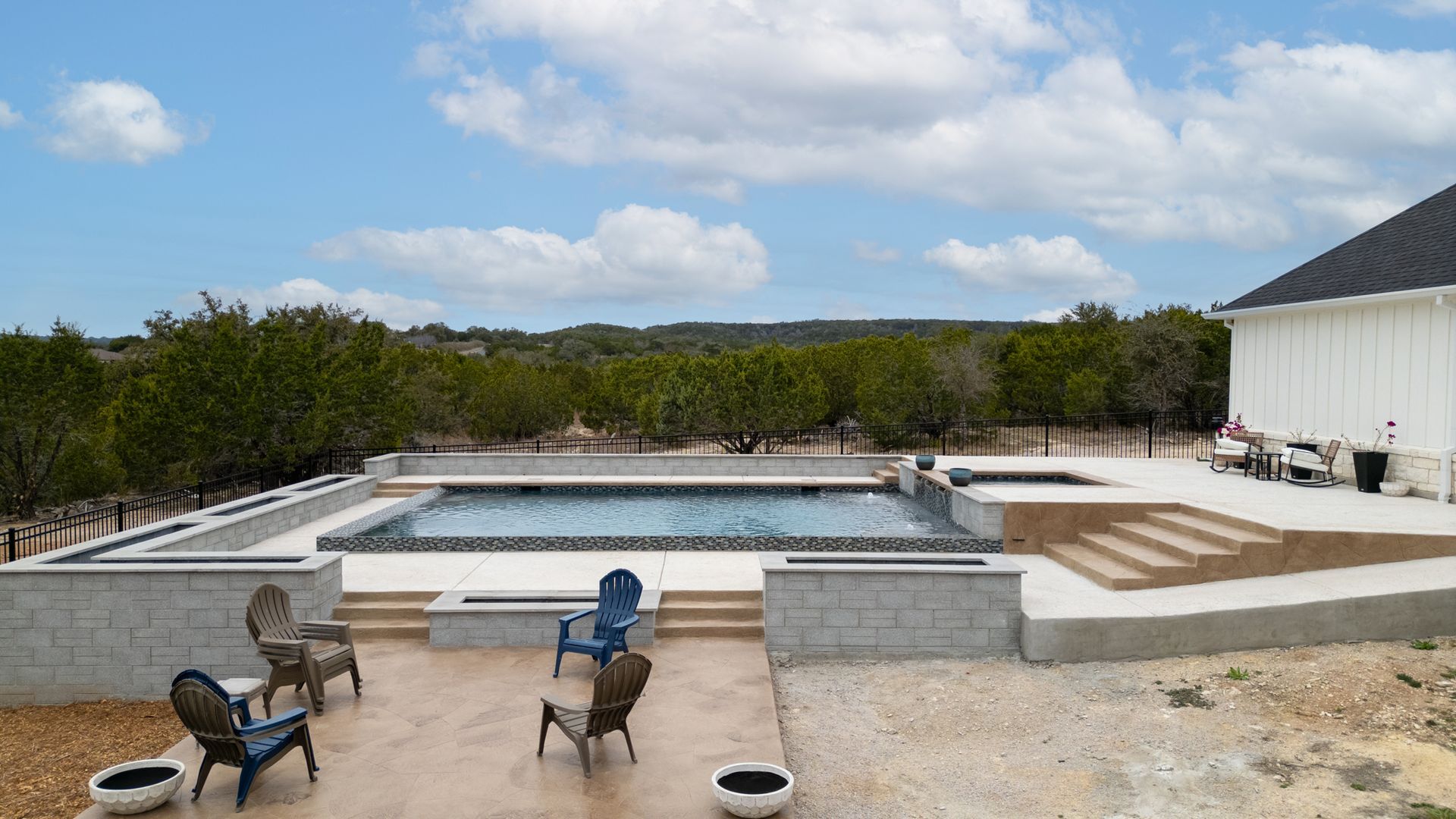 A pool area with a rectangular pool, stone patio, four outdoor chairs, and stairs leading to a white house under a blue sky.
