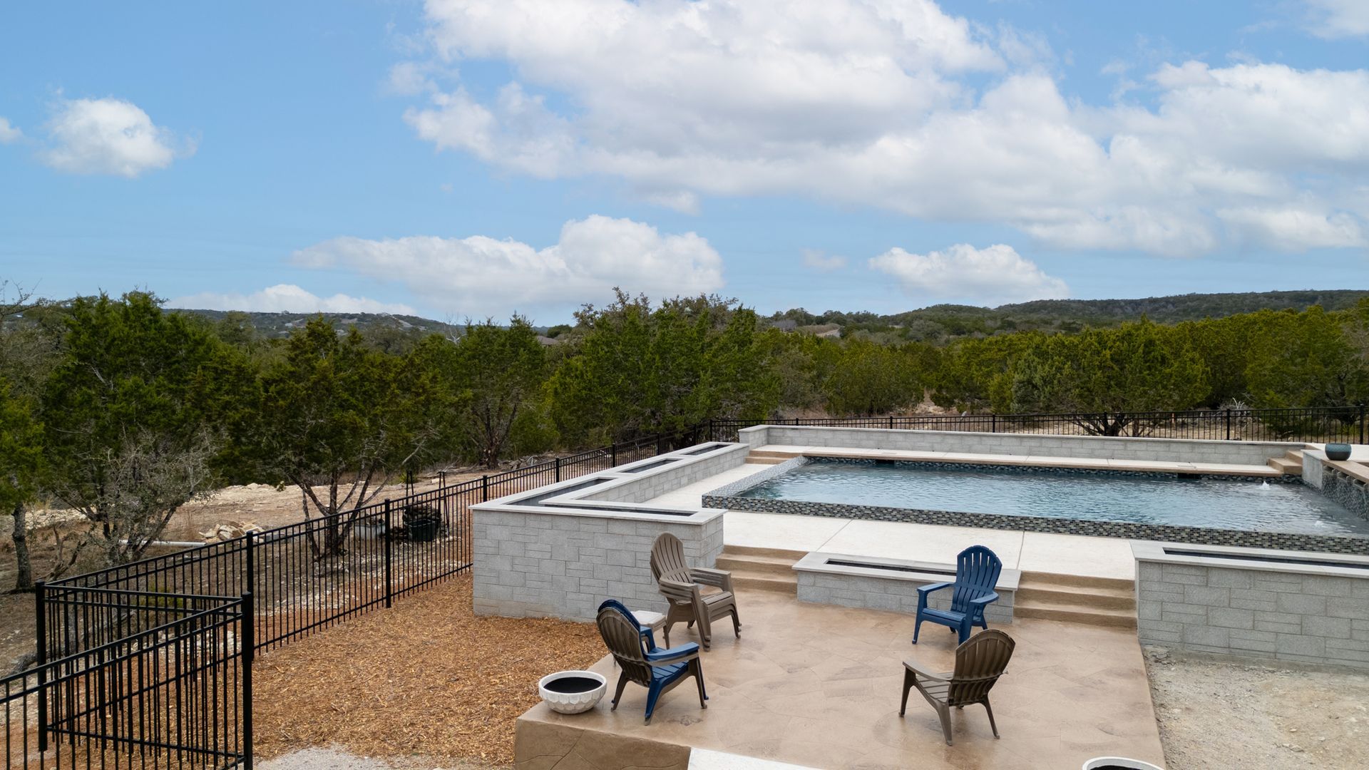 A rectangular swimming pool sits next to a patio with four outdoor chairs, surrounded by trees under a cloudy blue sky.