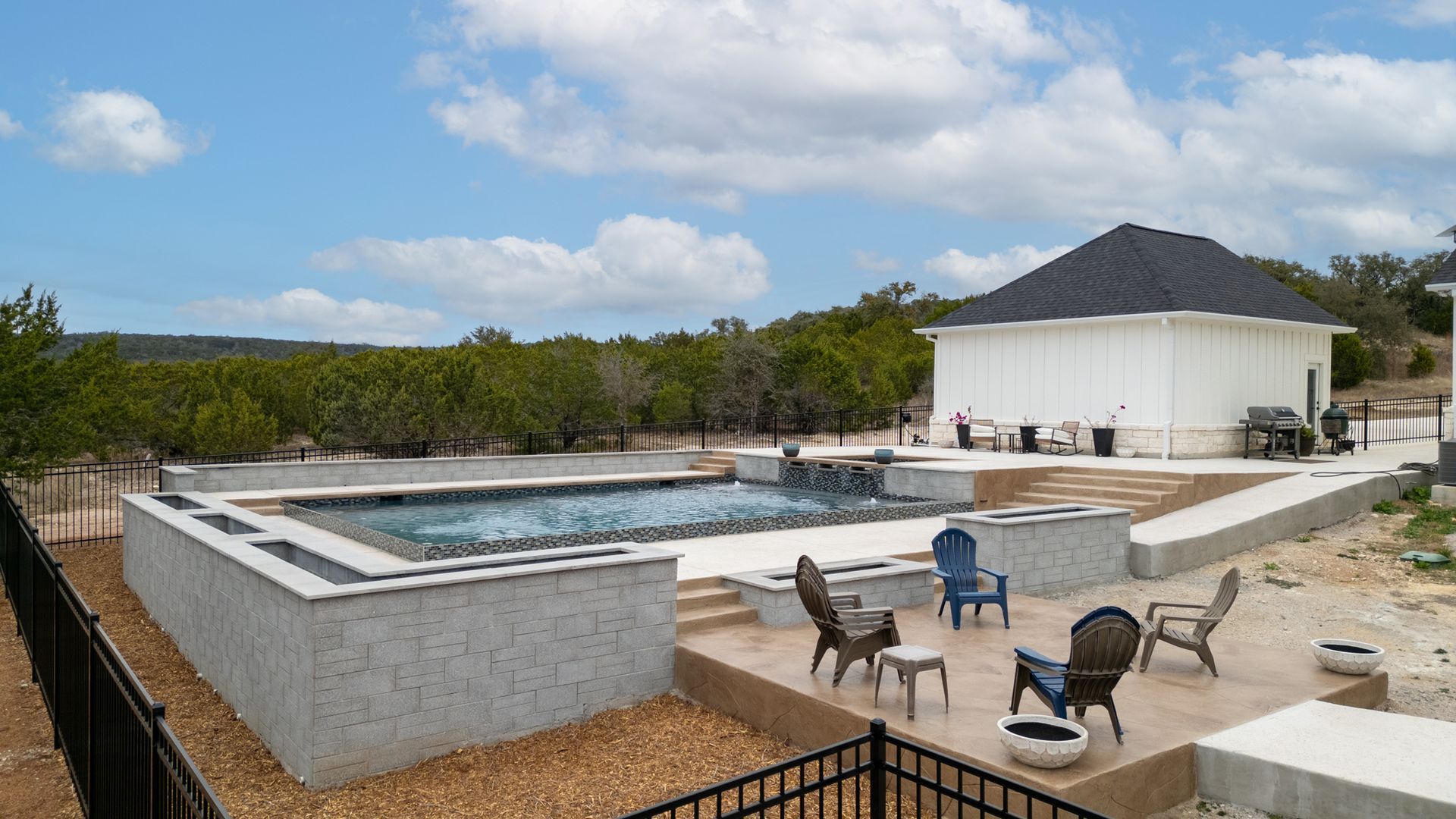 A backyard with a rectangular inground pool, stone patio, four outdoor chairs, and a white pool house under a blue sky.