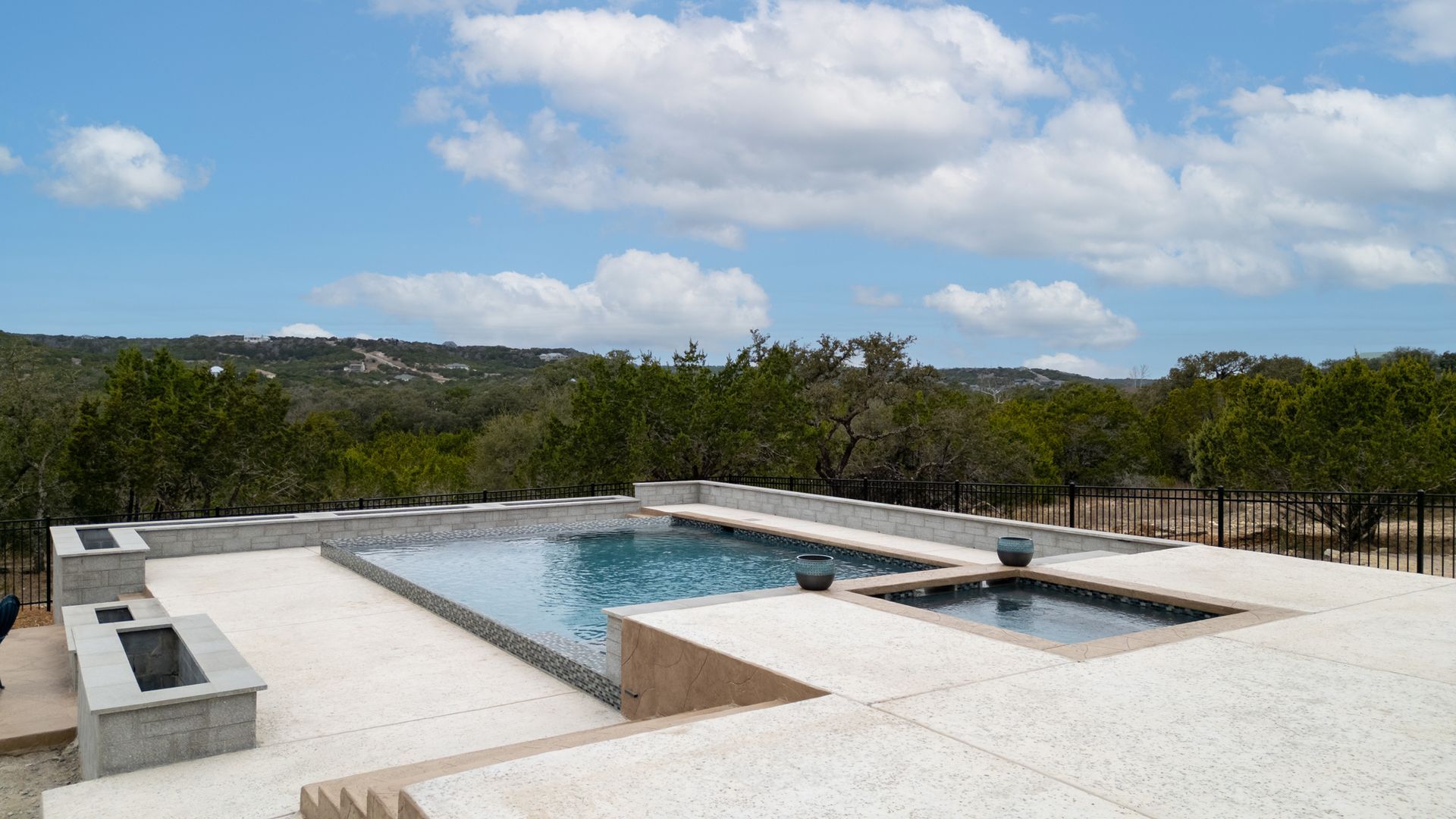 A rectangular swimming pool and a separate square spa on a stone patio overlooking a tree-lined landscape under a blue sky.