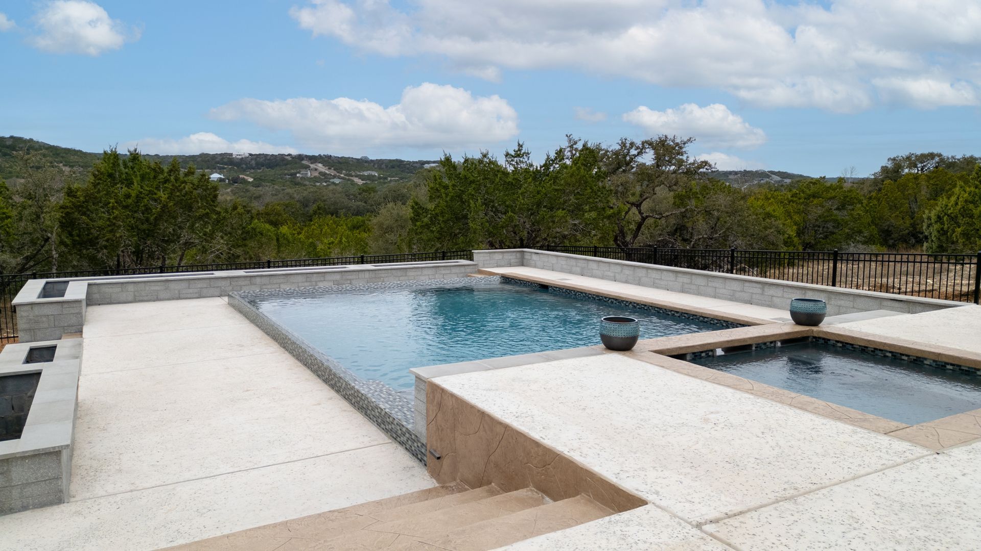 An outdoor swimming pool and attached hot tub on a concrete patio overlooking a wooded landscape under a cloudy sky.