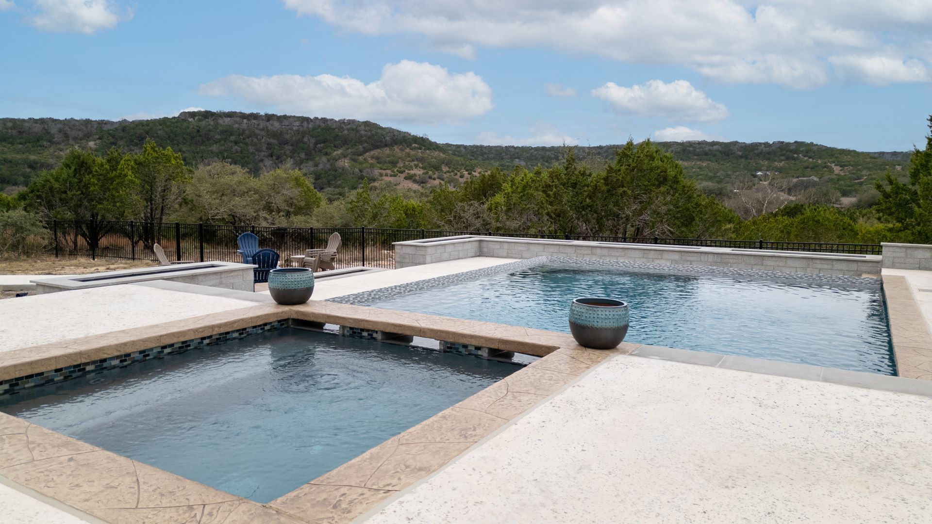 A split-level pool and hot tub set on a patio, overlooking a landscape of rolling hills and trees under a blue sky.