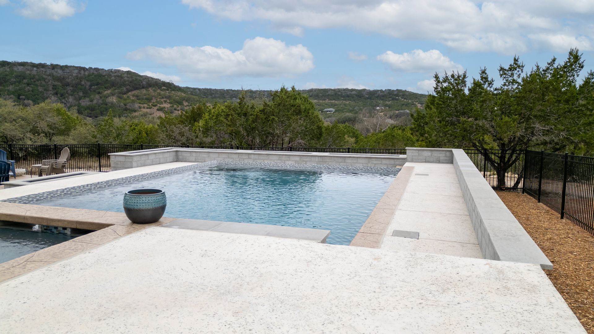 An infinity pool with light-colored decking and a basin centerpiece, overlooking a scenic landscape of hills and trees.